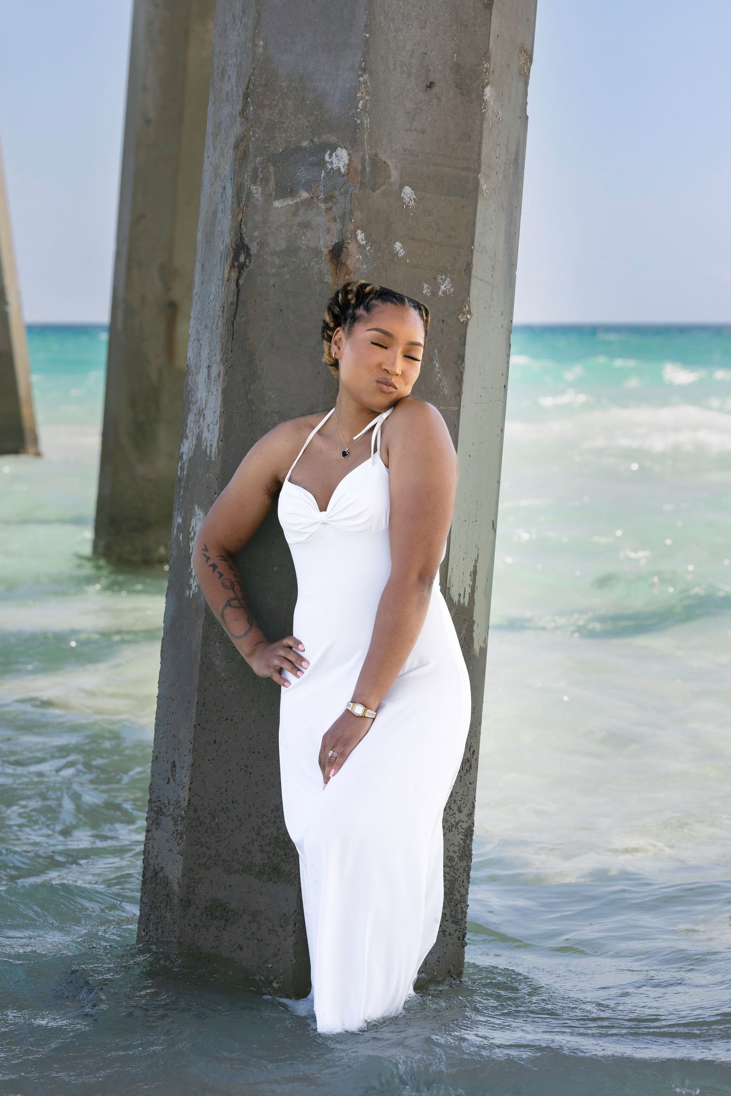 Woman in white maxi dress posing by concrete pier pillars in turquoise ocean water at a beach.