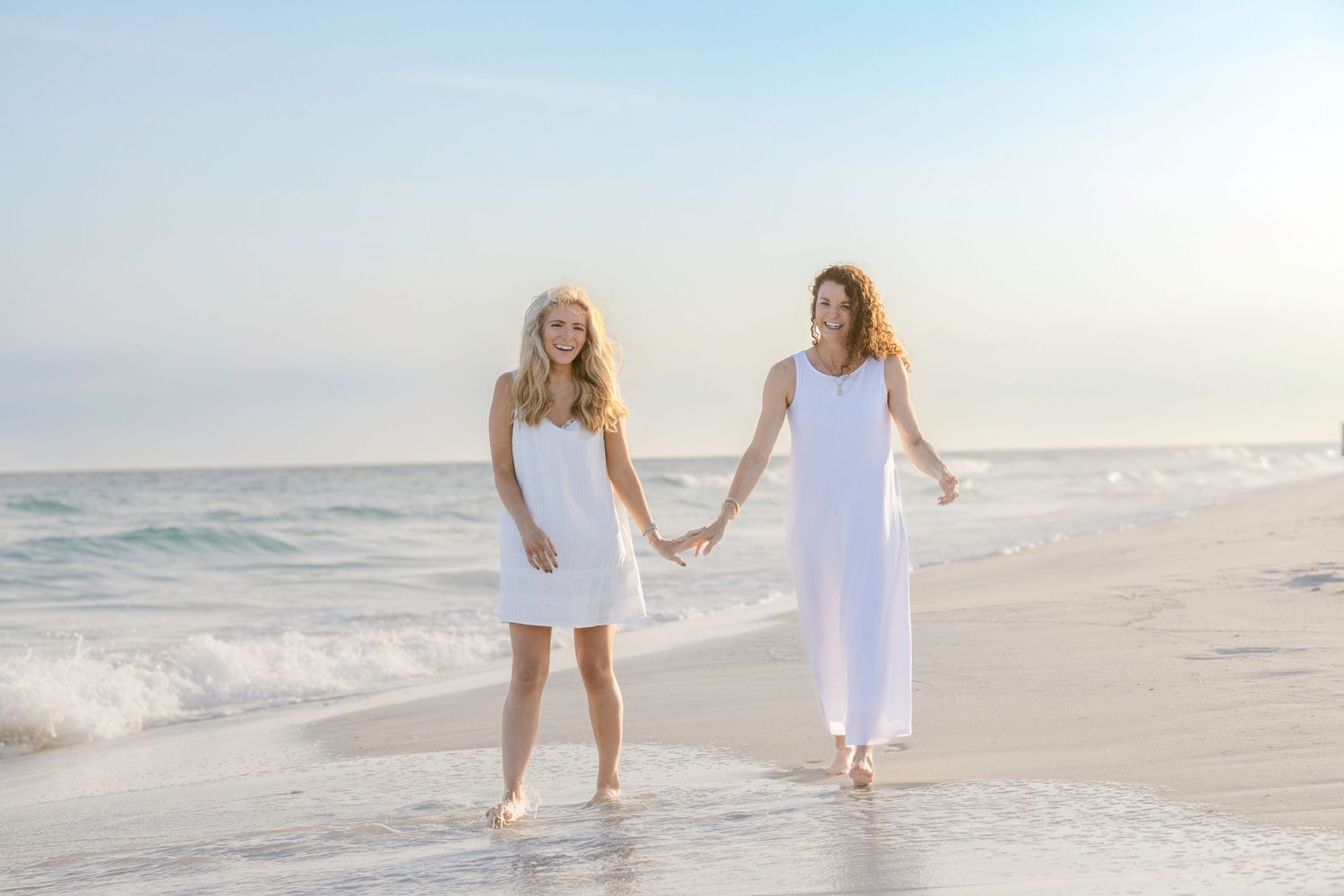 Two women in white dresses holding hands and walking barefoot on a sunny beach with gentle waves.