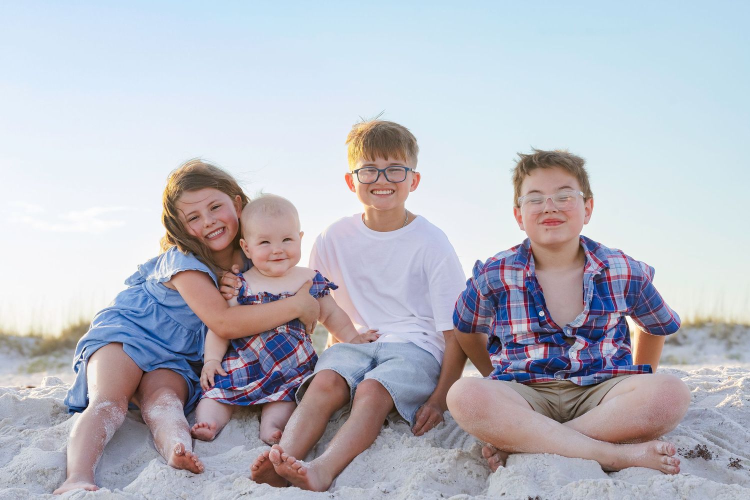 Four children sitting together on a sandy beach, smiling happily on a sunny day.