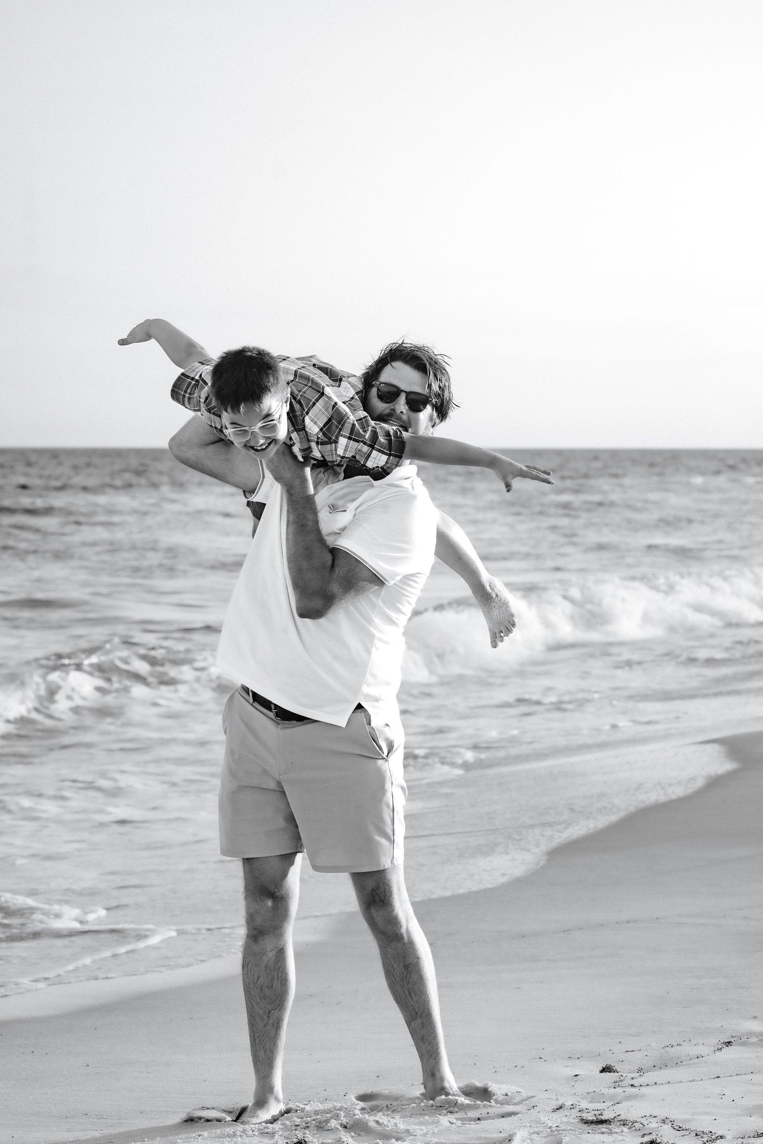 Father playfully lifting son on beach in black and white photo by the ocean waves.