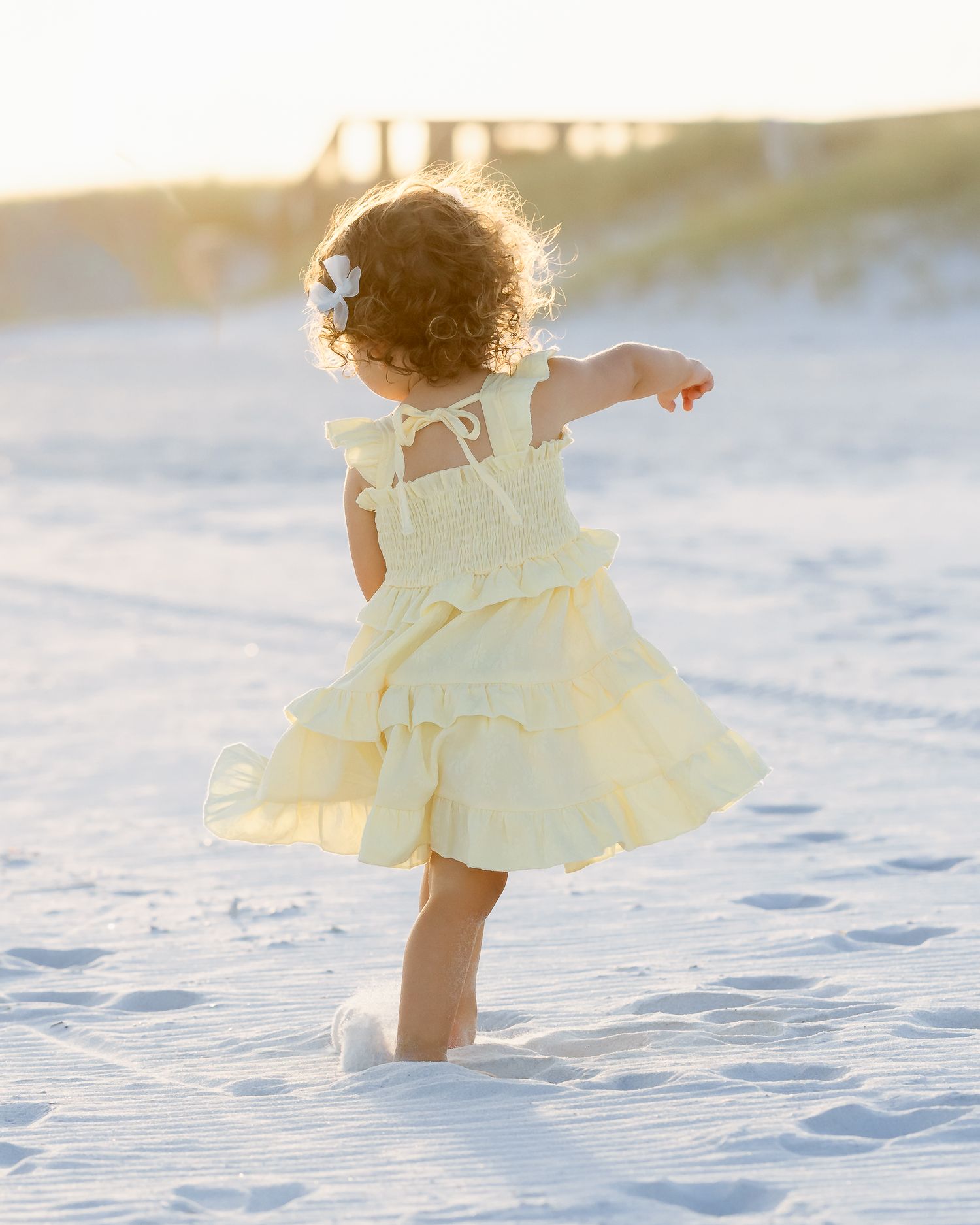Toddler girl in yellow ruffled dress walking on white sand beach at golden hour.