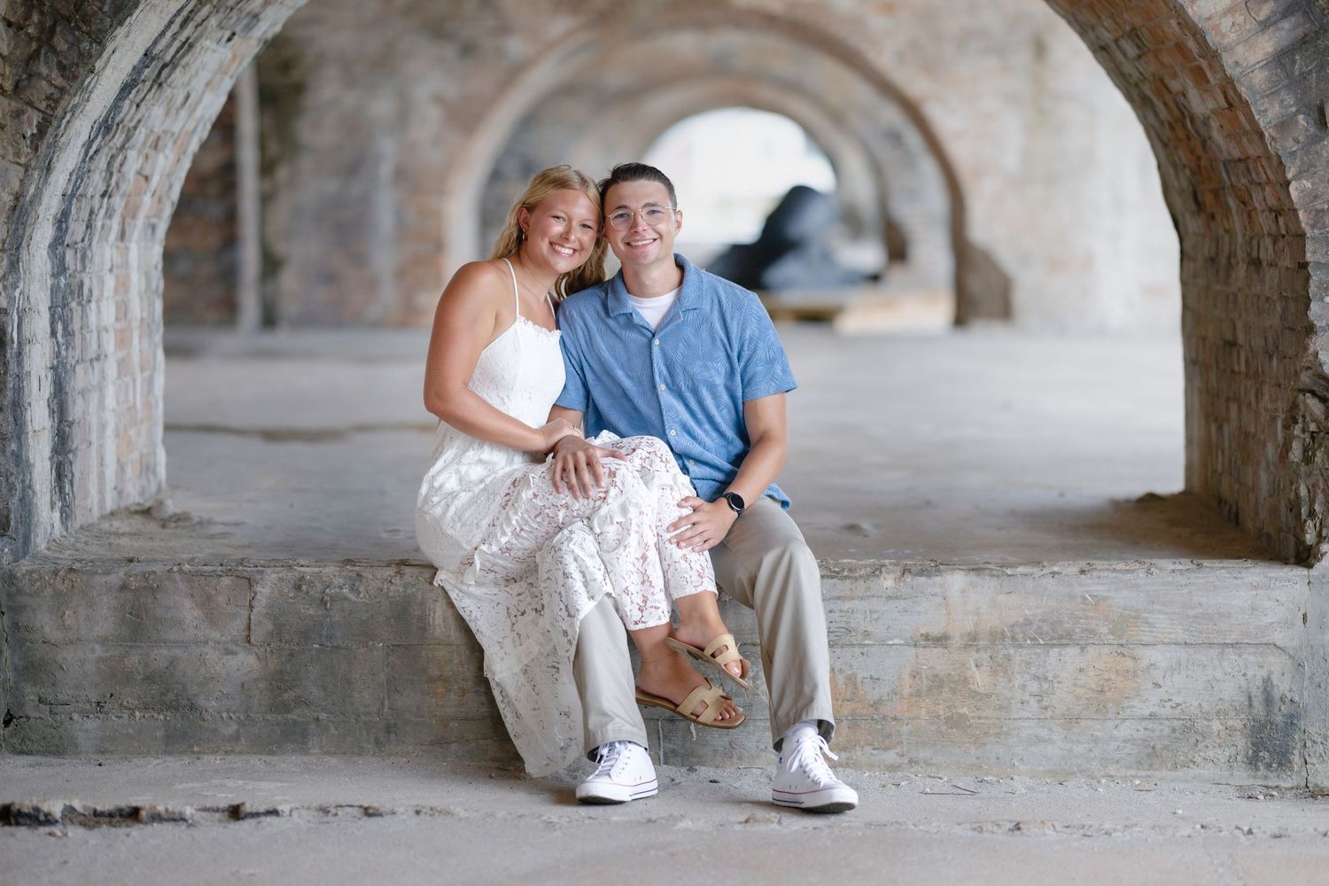 Couple sitting together under stone arches, smiling, woman in white dress, man in blue shirt, outdoor setting.