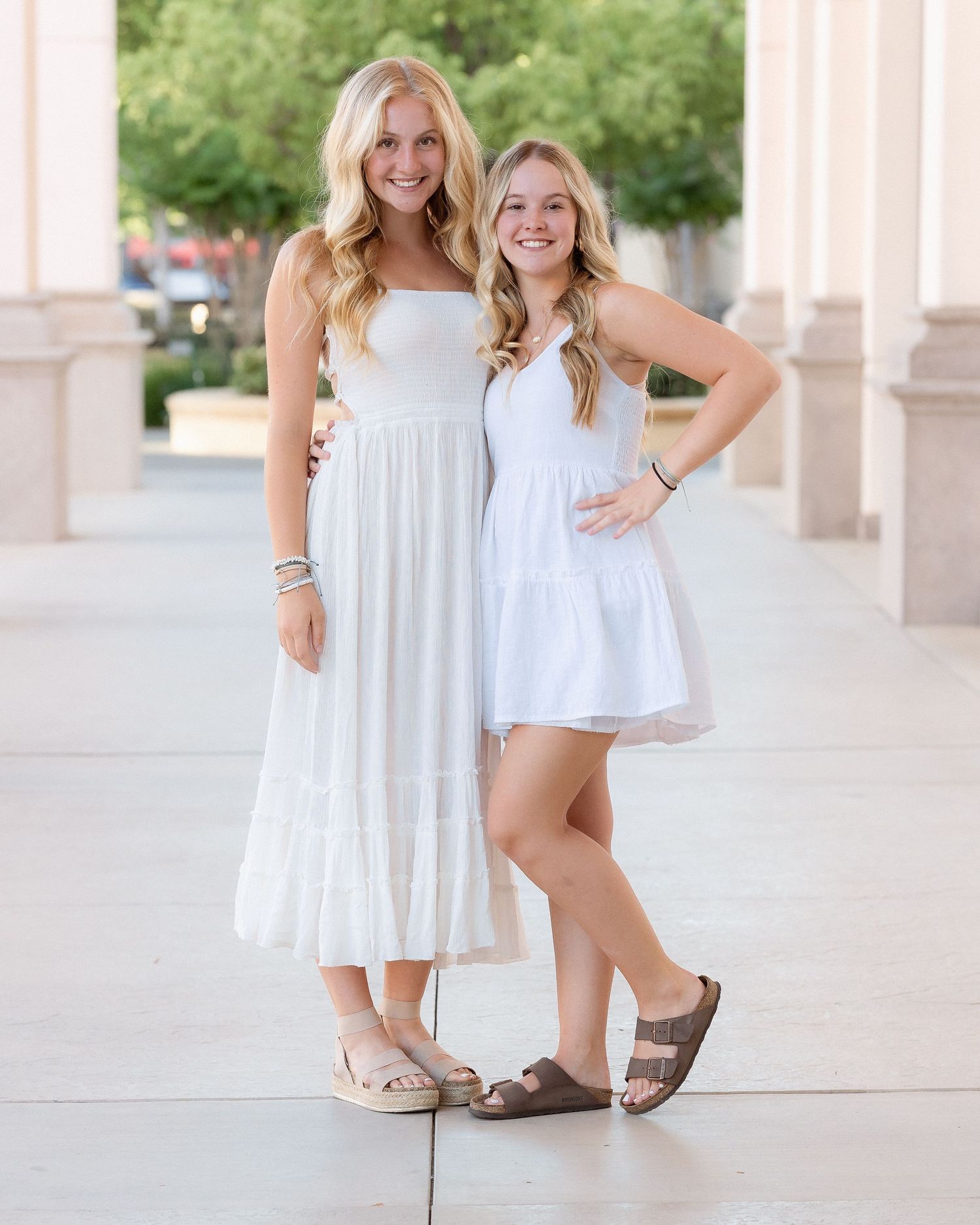 Two blonde sisters in white dresses smile together for a portrait between outdoor columns.