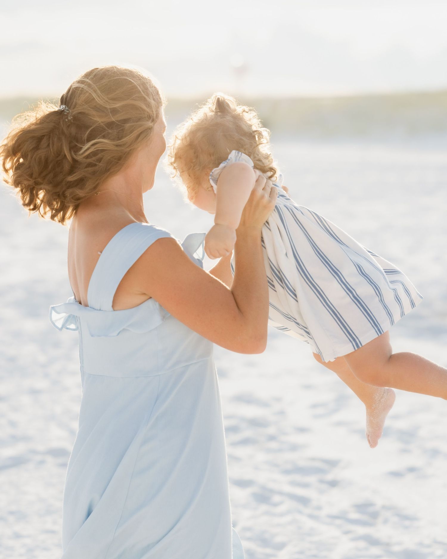 A mother lifts her toddler joyfully on a sunny beach, both wearing light blue outfits in soft golden light.