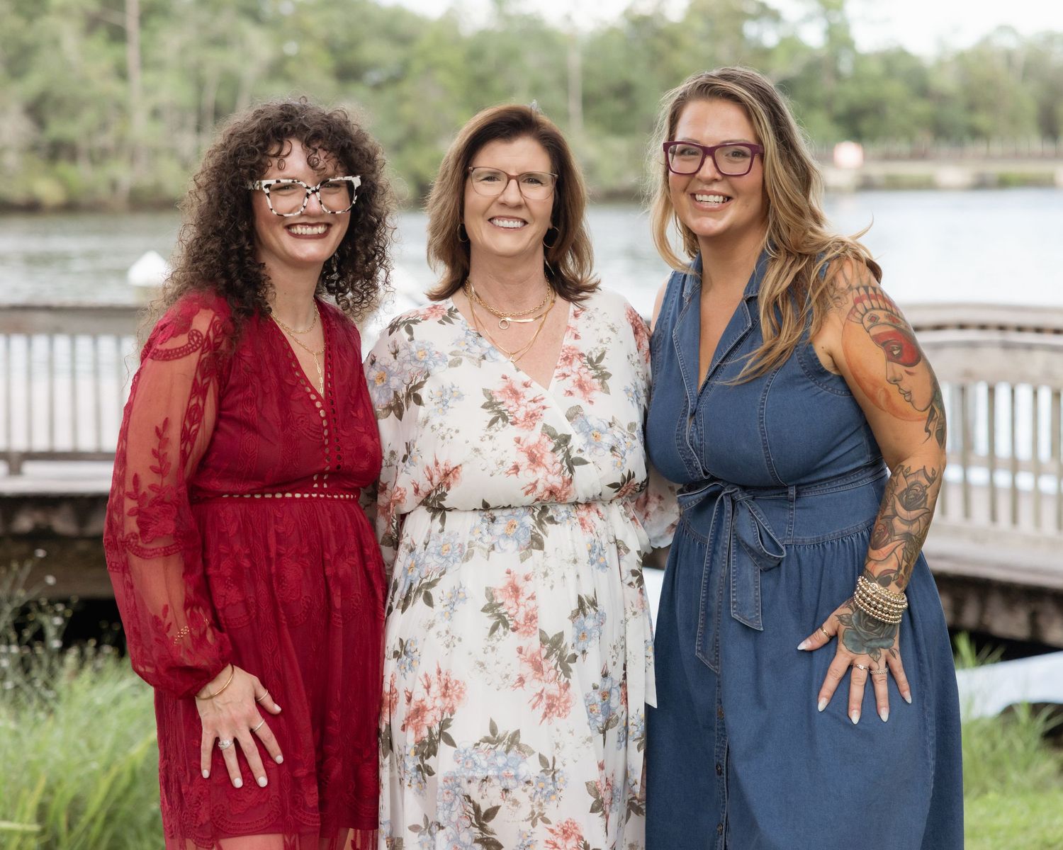 Three smiling women posing together in floral and solid dresses near a scenic waterfront.