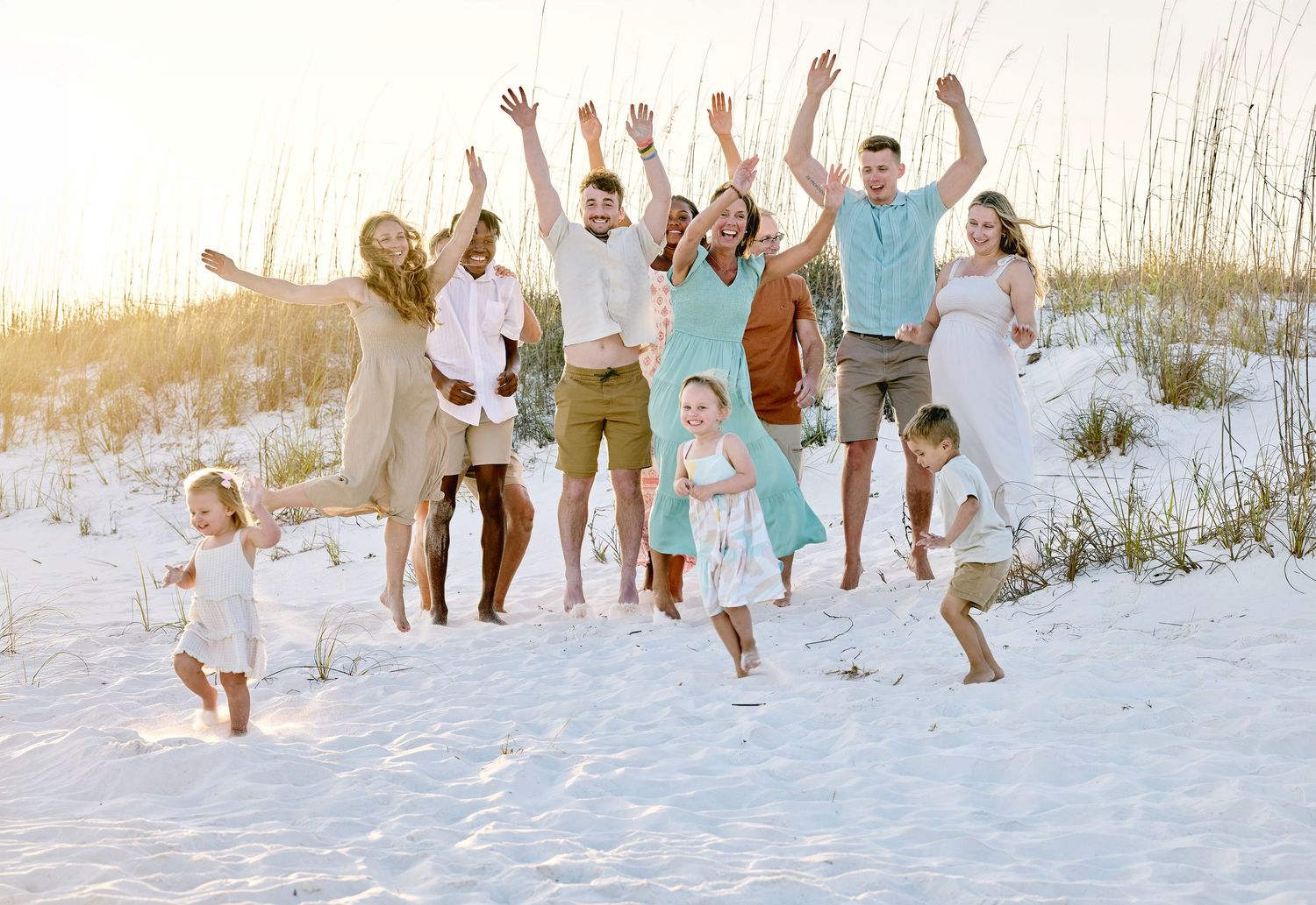 Large family group jumping joyfully on white sand beach at sunset, wearing coordinated outfits.