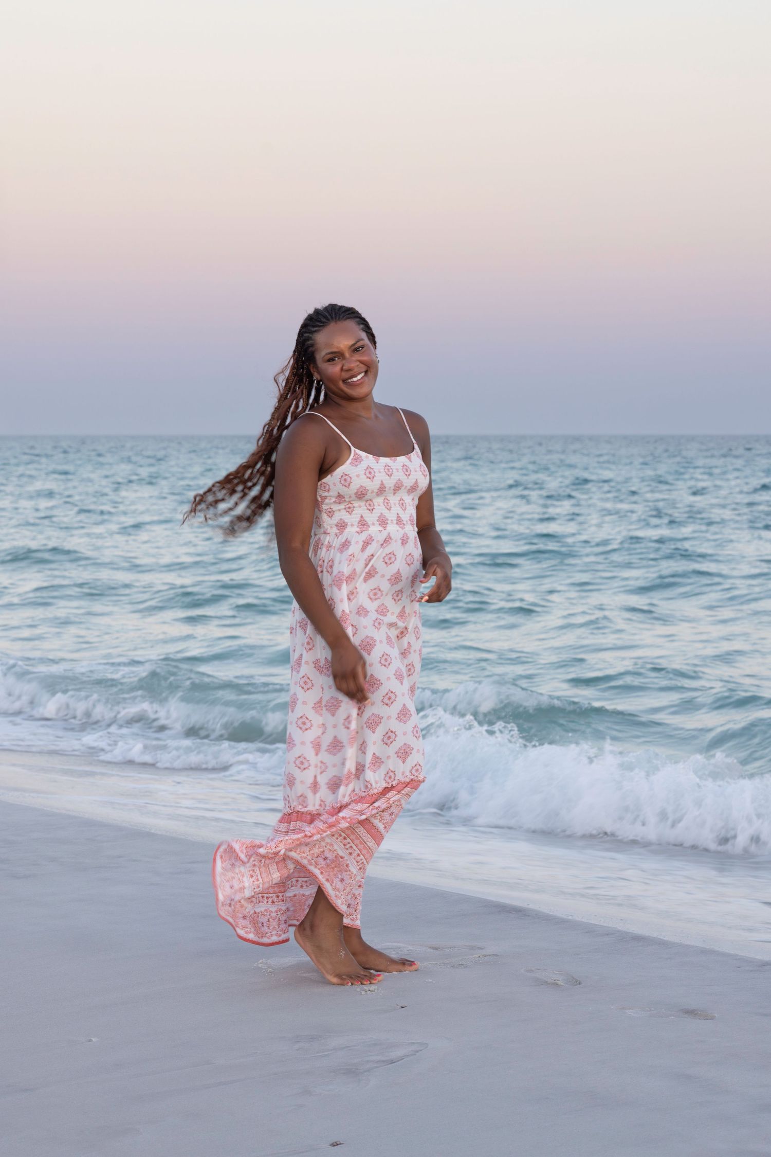 A woman in a floral maxi dress walks barefoot along a sandy beach at dusk.