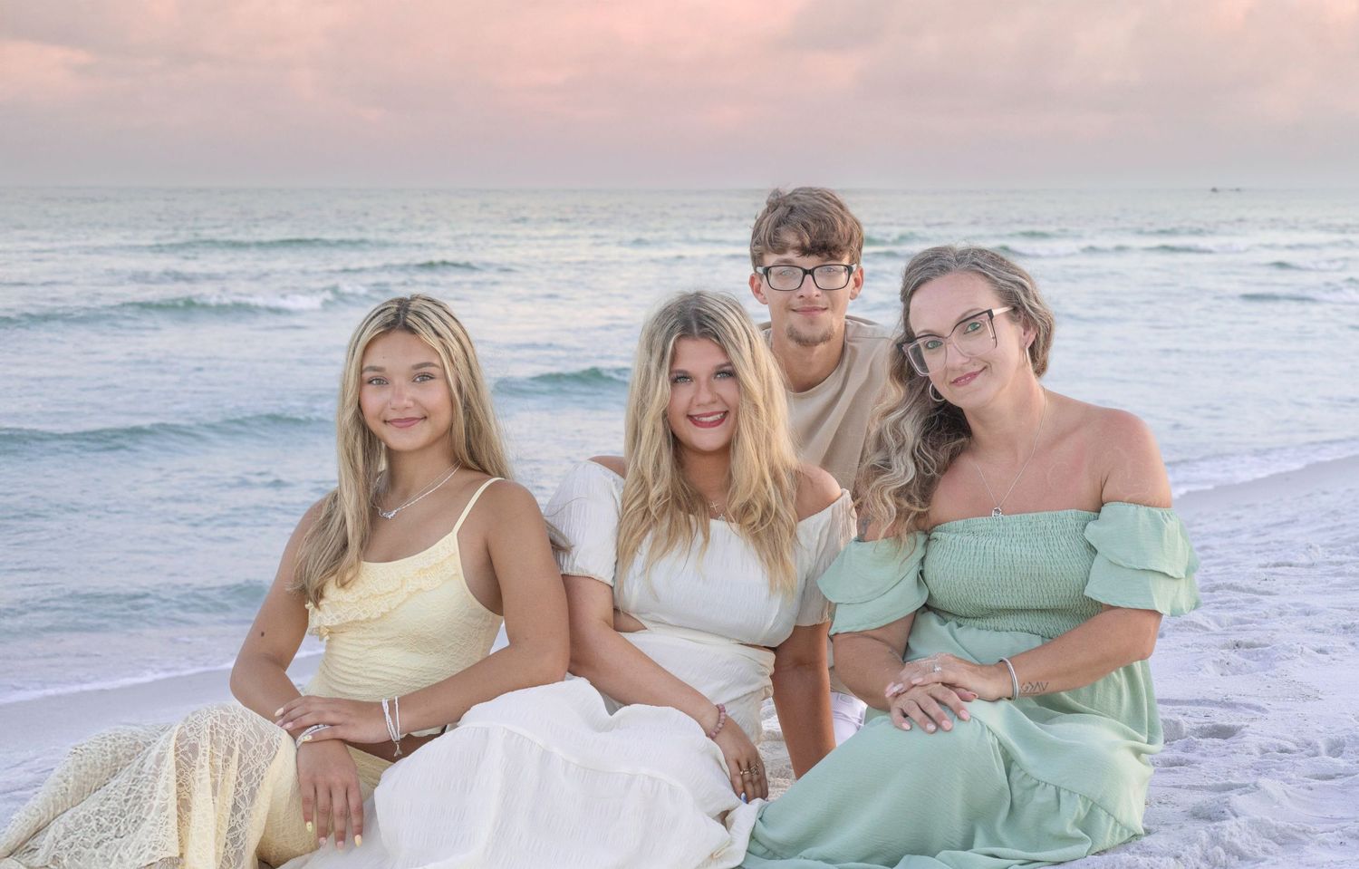 A group of friends in soft pastel outfits pose together at the beach during sunset, creating a dreamy coastal scene.