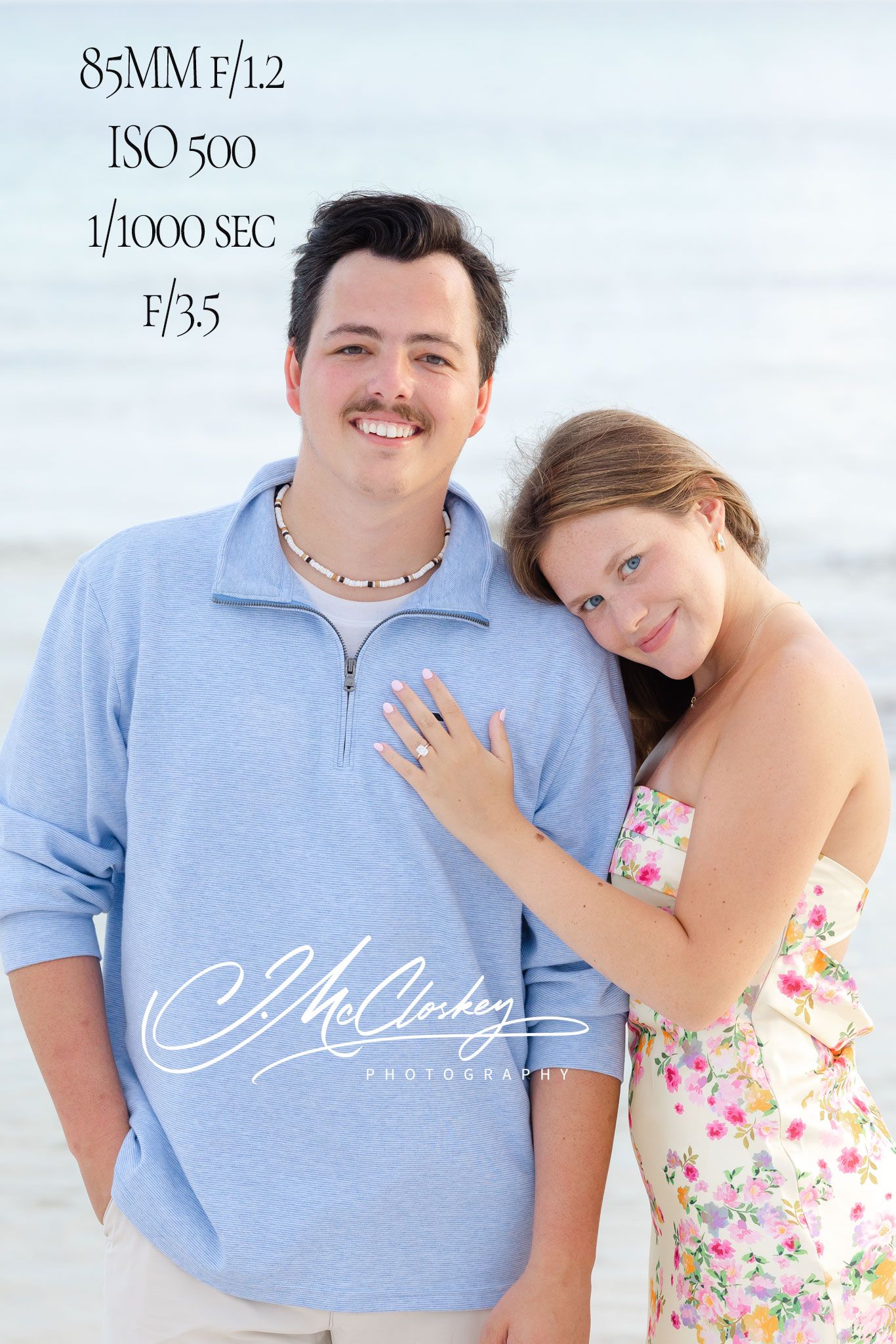 A couple poses together on a beach, with camera settings displayed at the top showing 85MM f/1.2 ISO 500 specs.