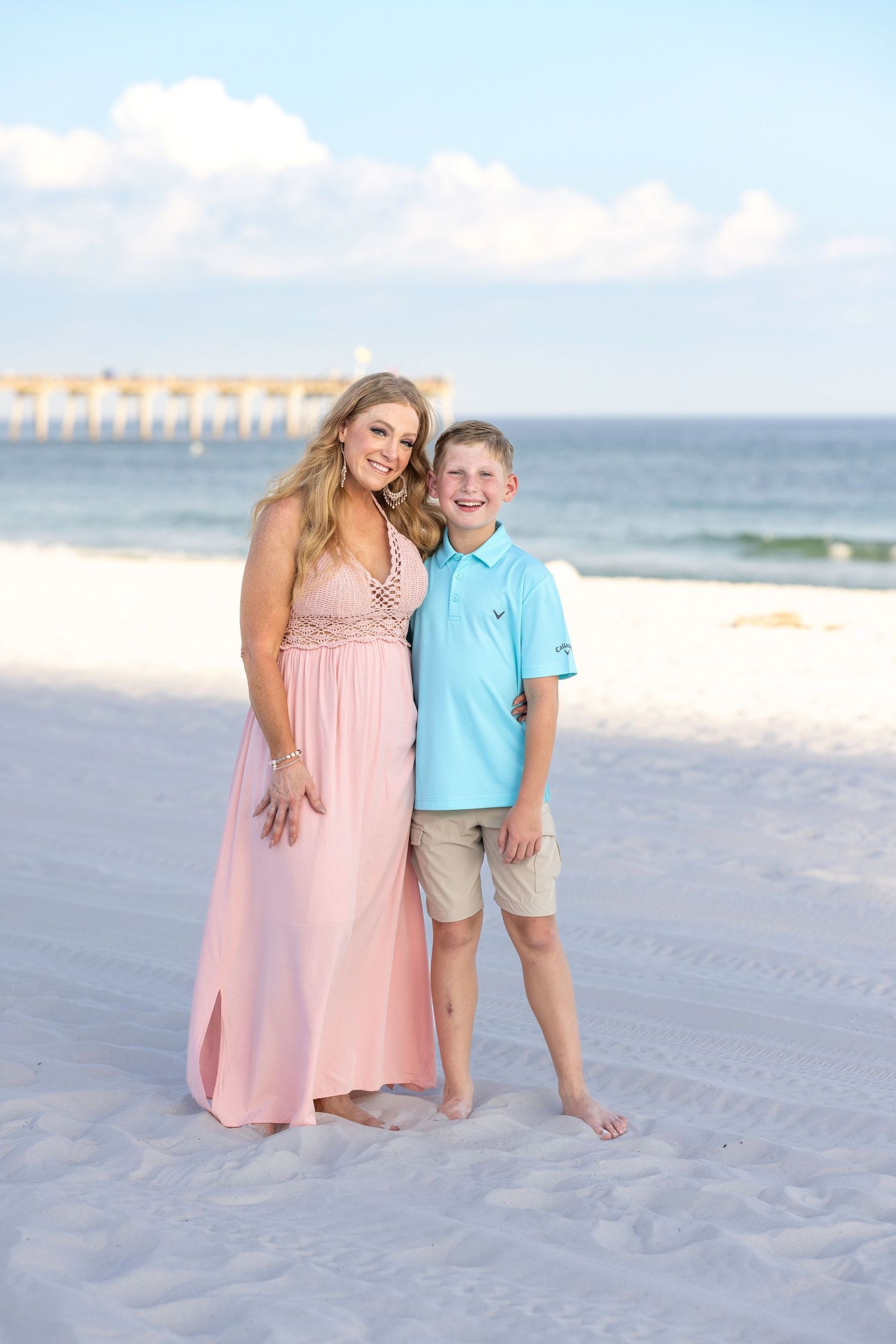 Two people in coordinating outfits pose together on a beautiful white sand beach with ocean waves in the background.
