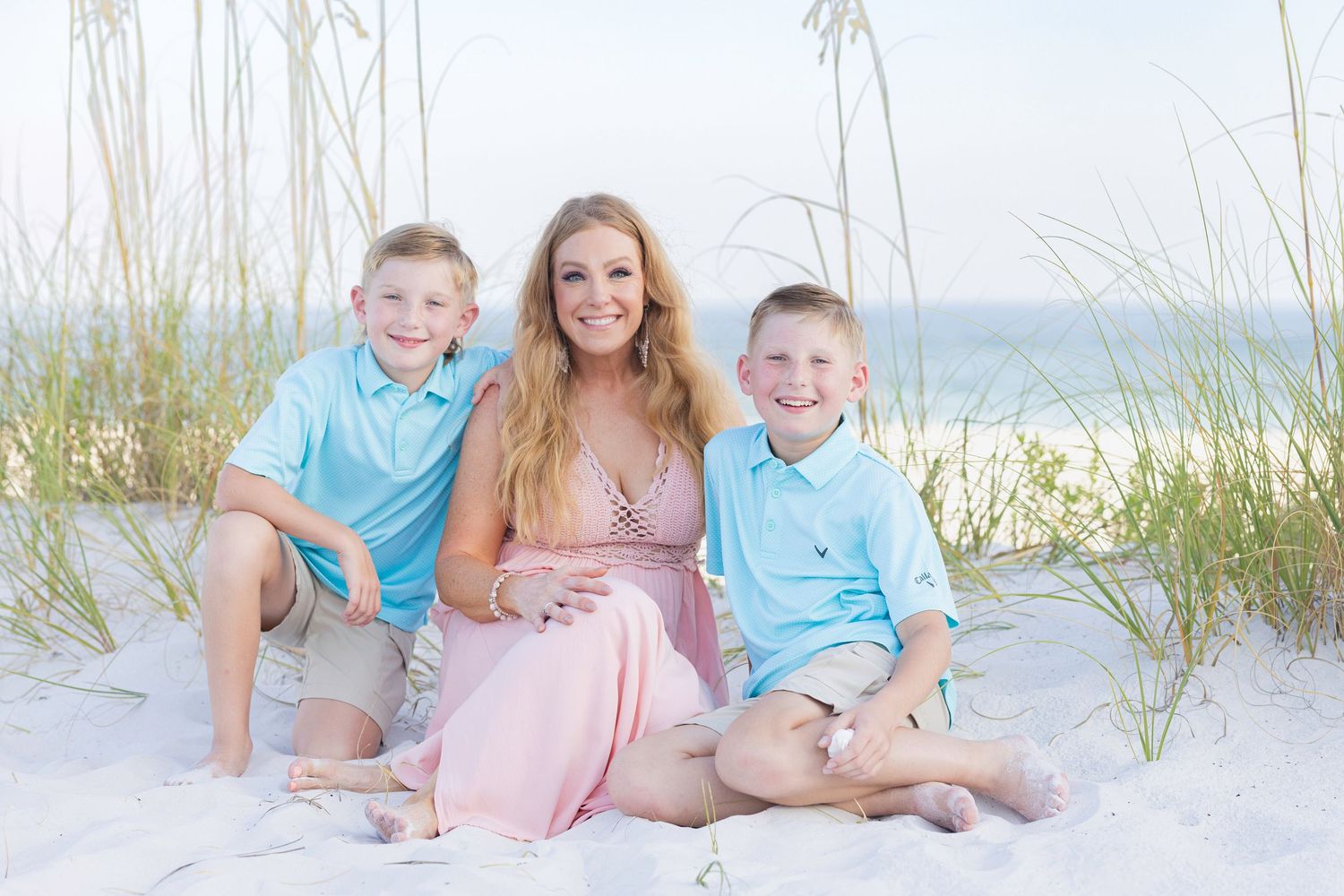 A beach portrait featuring matching turquoise outfits and a pink dress set against white sand dunes and beach grass.