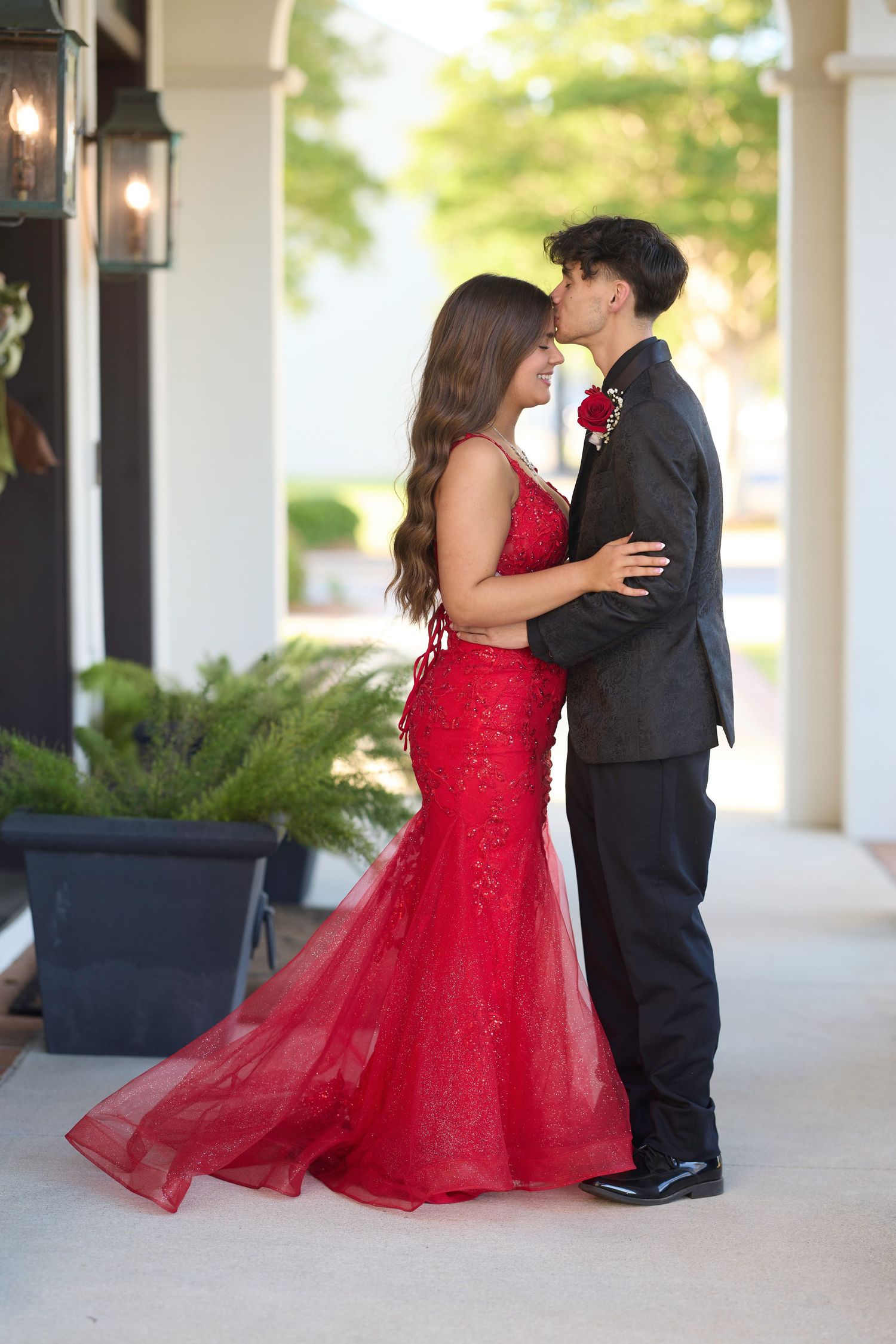 A couple in formal attire embrace during what appears to be prom, with the woman wearing a stunning red gown.