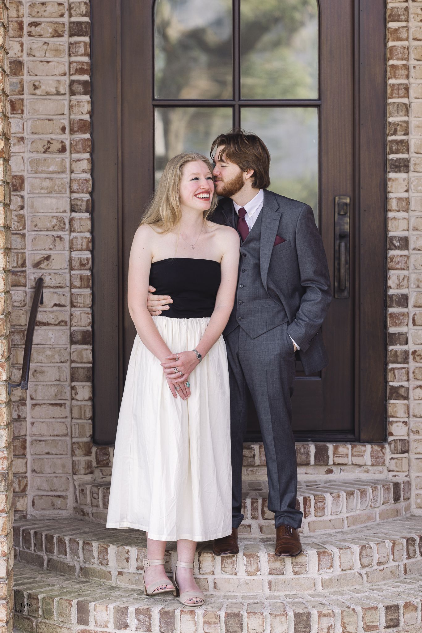 A couple shares a sweet moment on brick steps in front of a glass door, wearing formal black and white attire.
