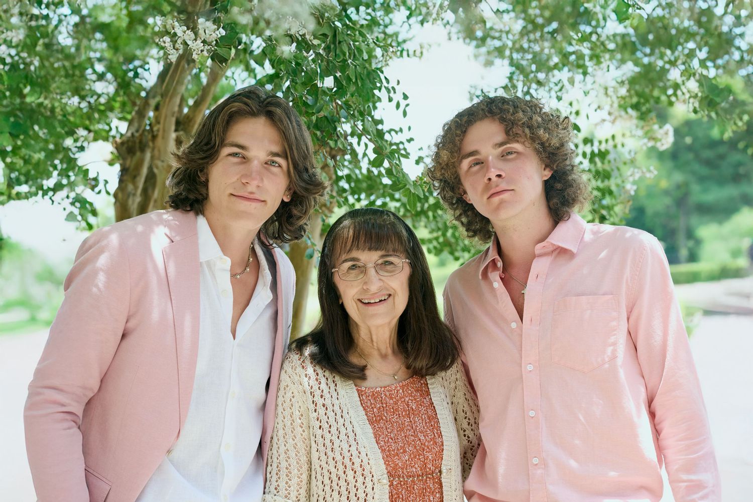 Two young men in pink shirts pose with an elderly woman outdoors under a tree in a warm family portrait.