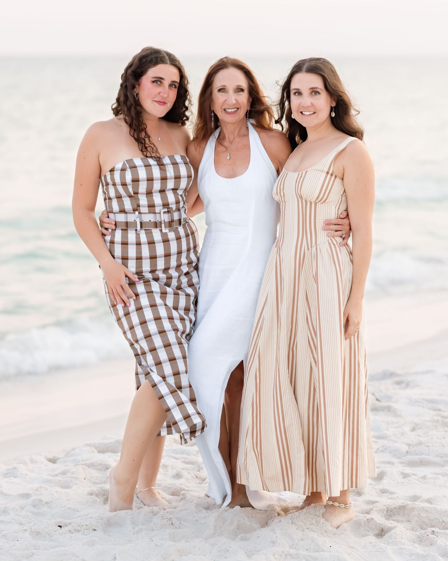 Three women in summer dresses pose together smiling on a sandy beach with ocean waves in the background.