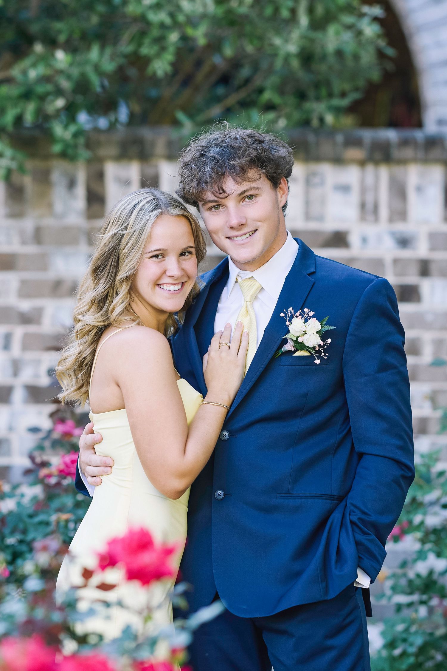 A young couple poses for a formal photo outdoors, with the woman in yellow and man in navy suit against stone steps.