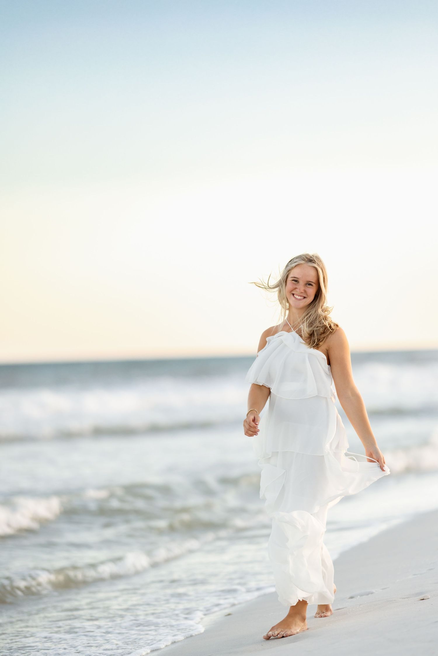Smiling blonde woman in flowing white maxi dress walks barefoot along shoreline with ocean waves behind her.