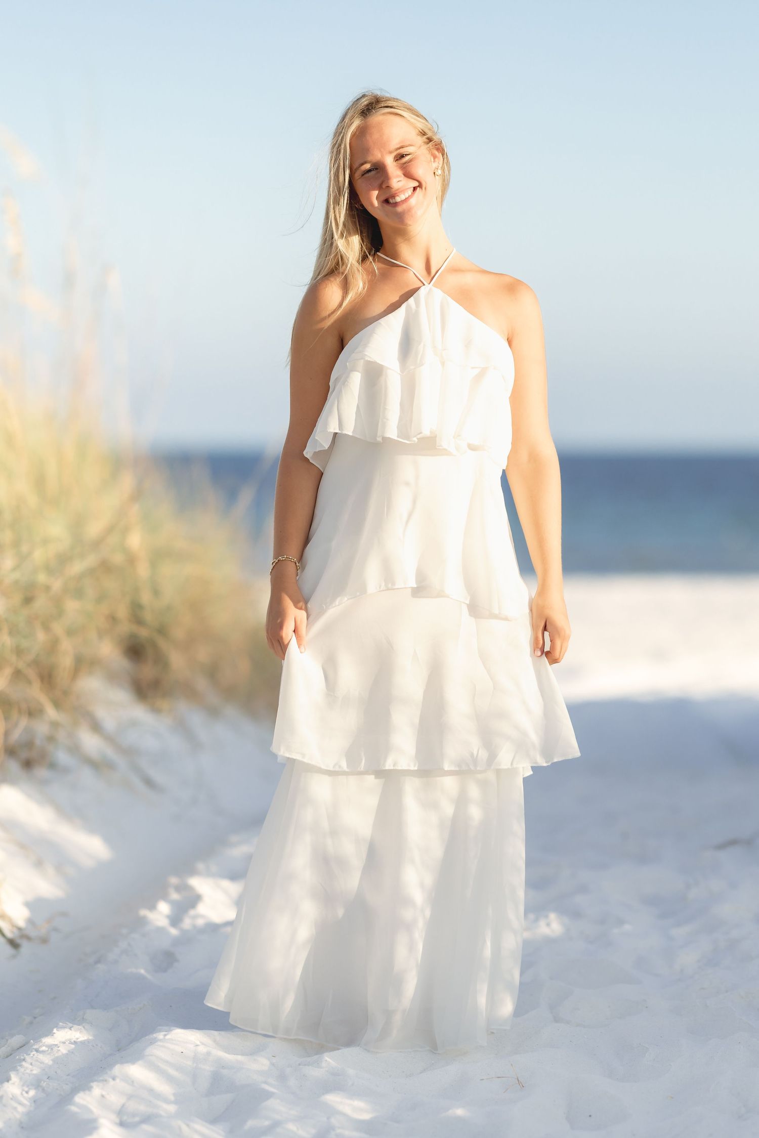 Happy blonde woman in white layered ruffle halter maxi dress laughing on sunny beach with ocean.