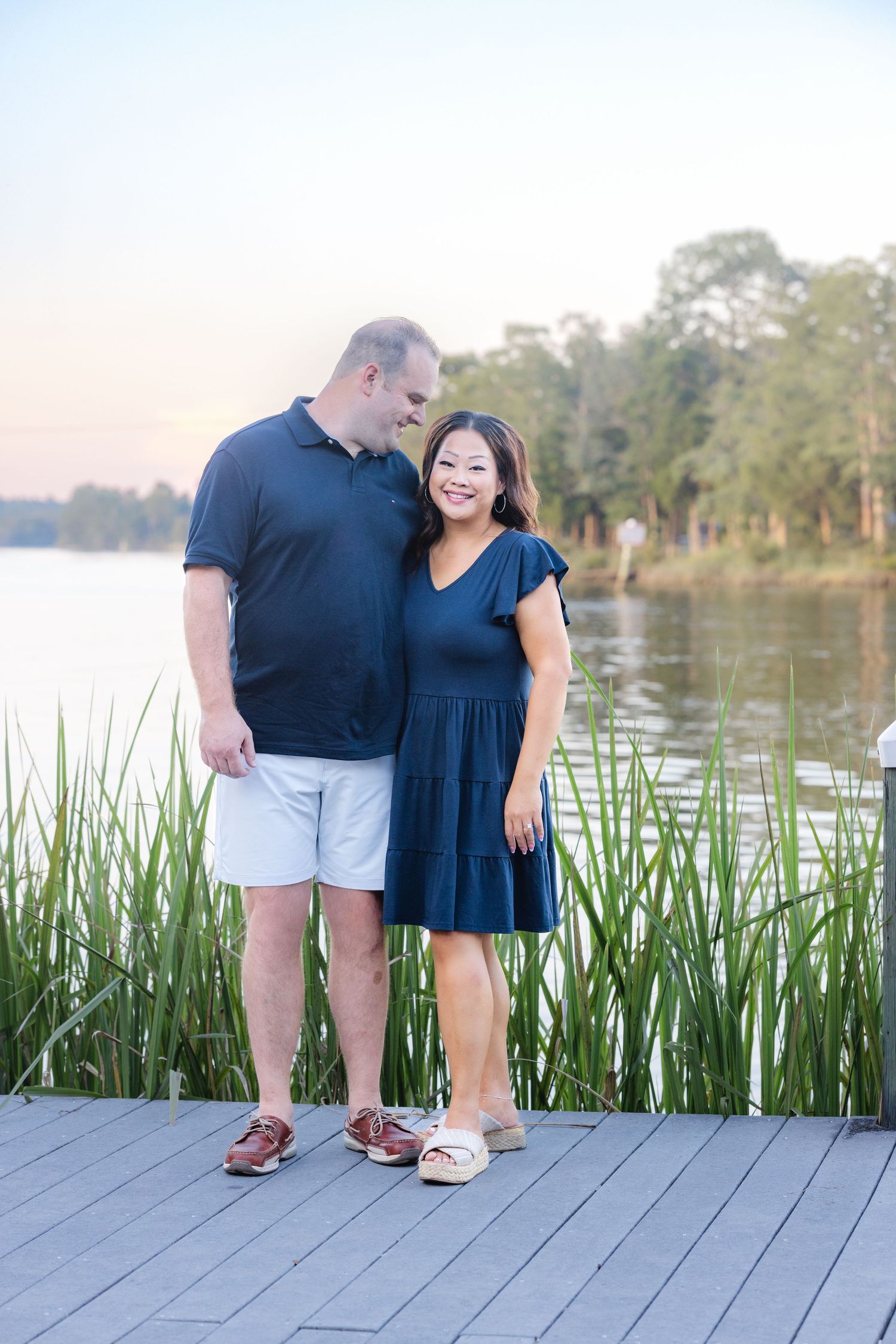 Couple in matching navy outfits smiling on a lakeside dock surrounded by tall grass at sunset.