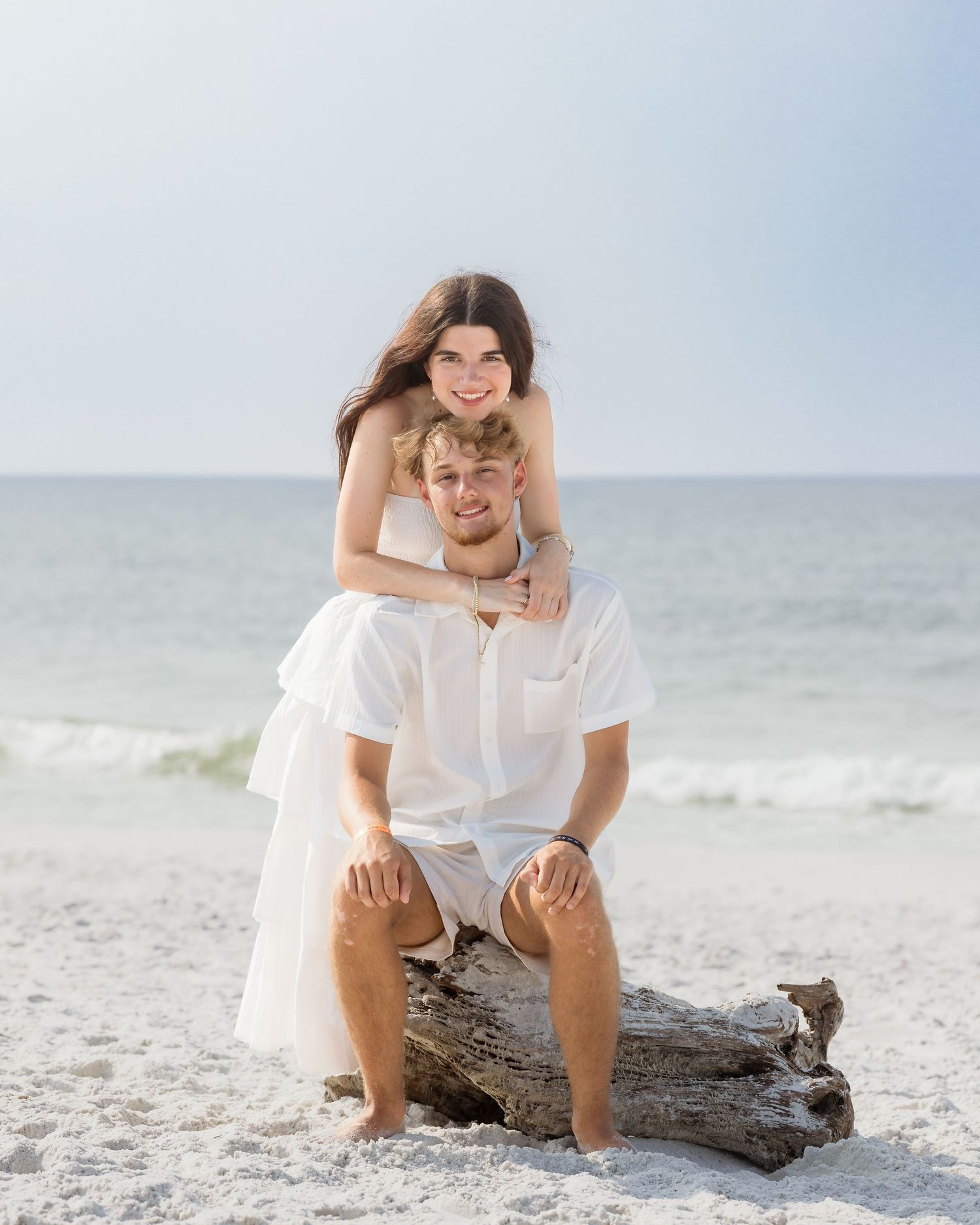Couple posing on driftwood at the beach, wearing white outfits, with the ocean in the background.