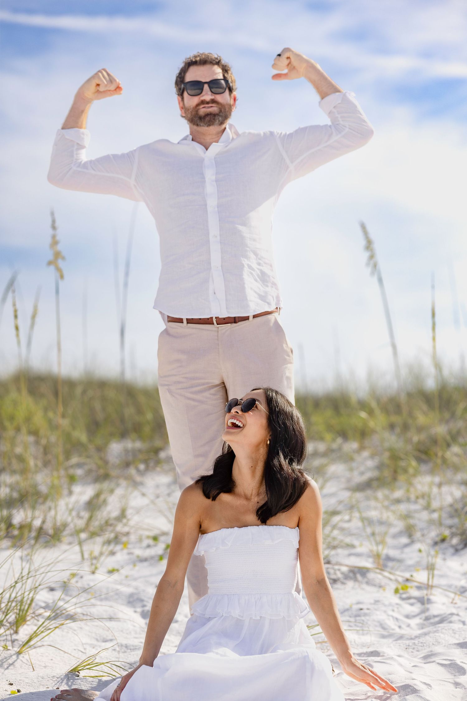 Happy couple on sandy beach, man flexing arms in white shirt, woman in white dress laughing, sunny day.