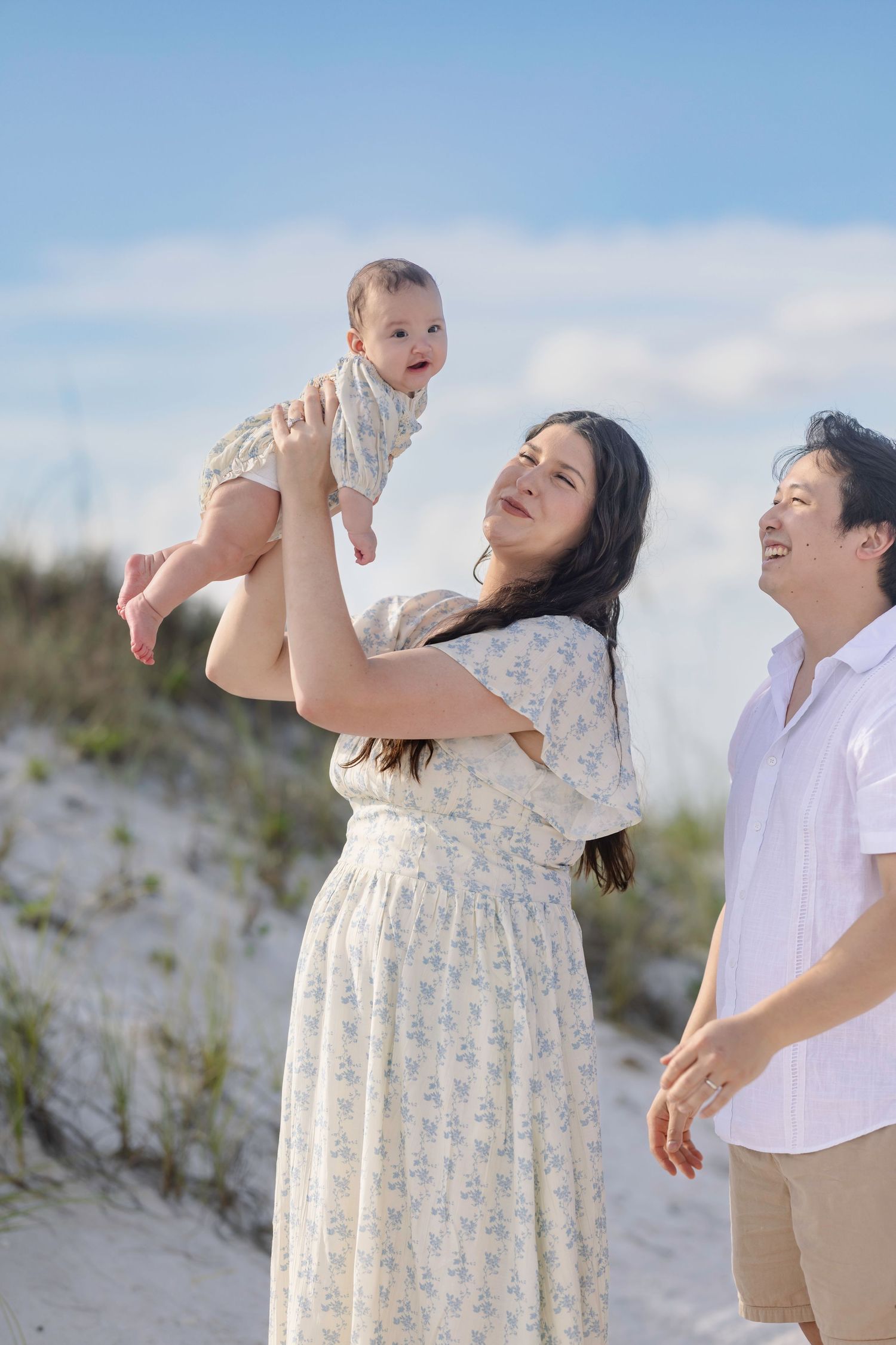 A joyful family on a beach, mother lifting baby in the air while father smiles, with sandy dunes and blue sky behind.