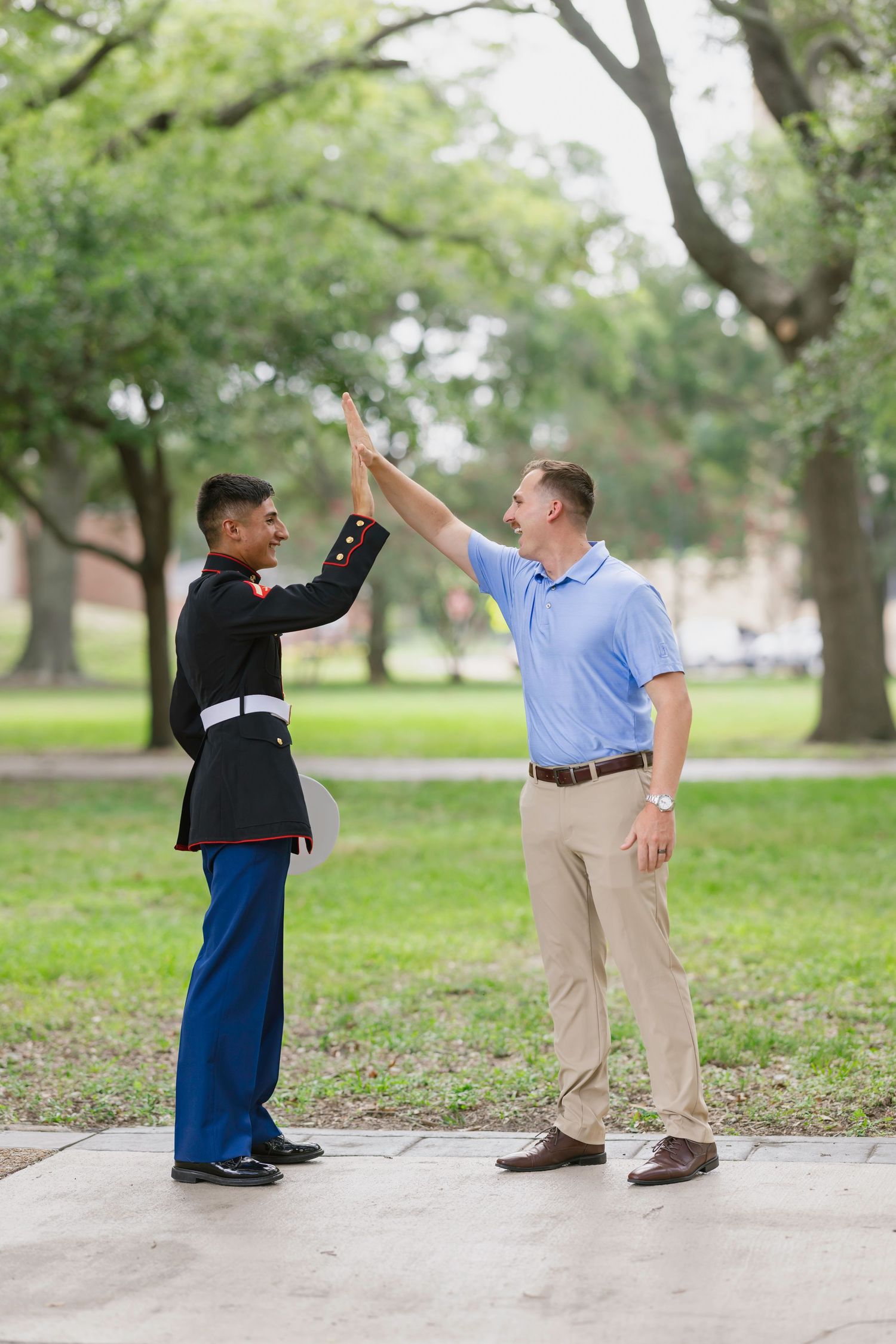 Marine in dress uniform gives high-five to man in blue polo shirt outdoors in green park setting.