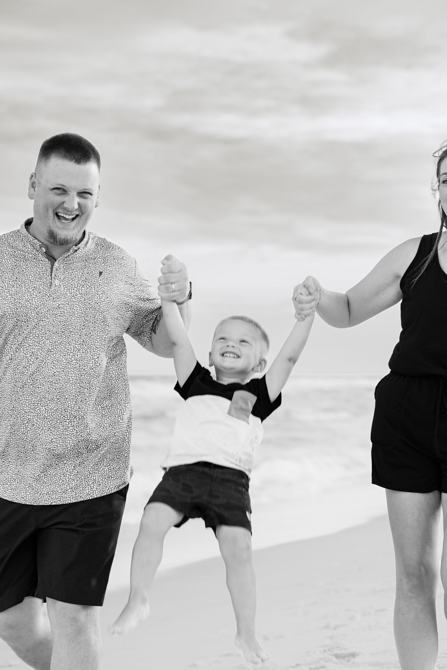 Black and white photo of happy family at beach, parents swinging young child by hands, smiling outdoors.
