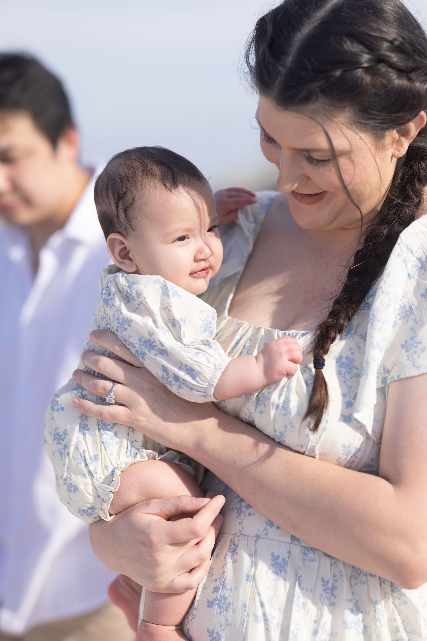 A mother with a dark braid holds her smiling baby outdoors, both dressed in floral outfits, with a man visible behind them.