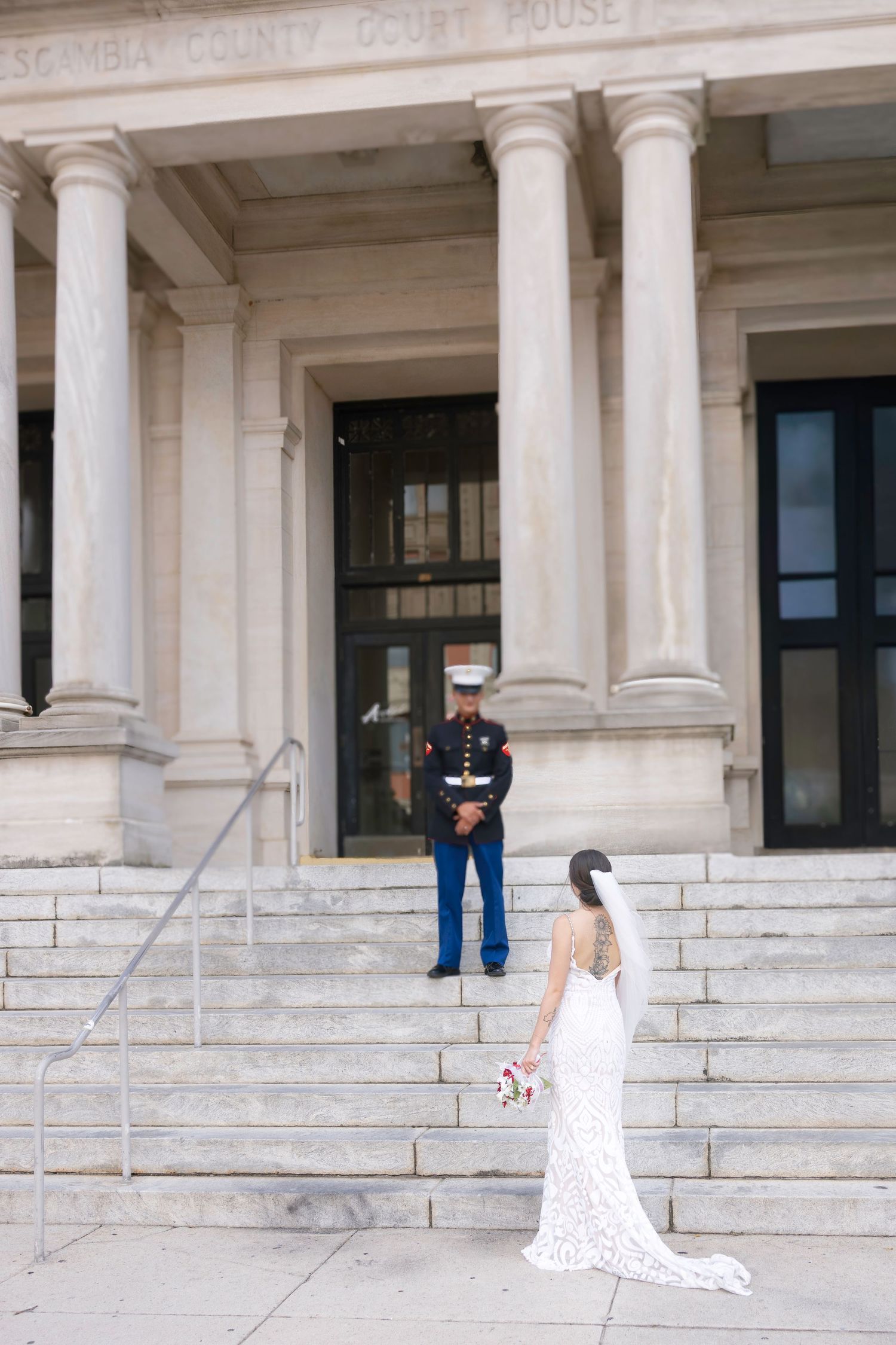 Marine in dress blues stands at courthouse steps as bride in white gown approaches for their wedding ceremony.