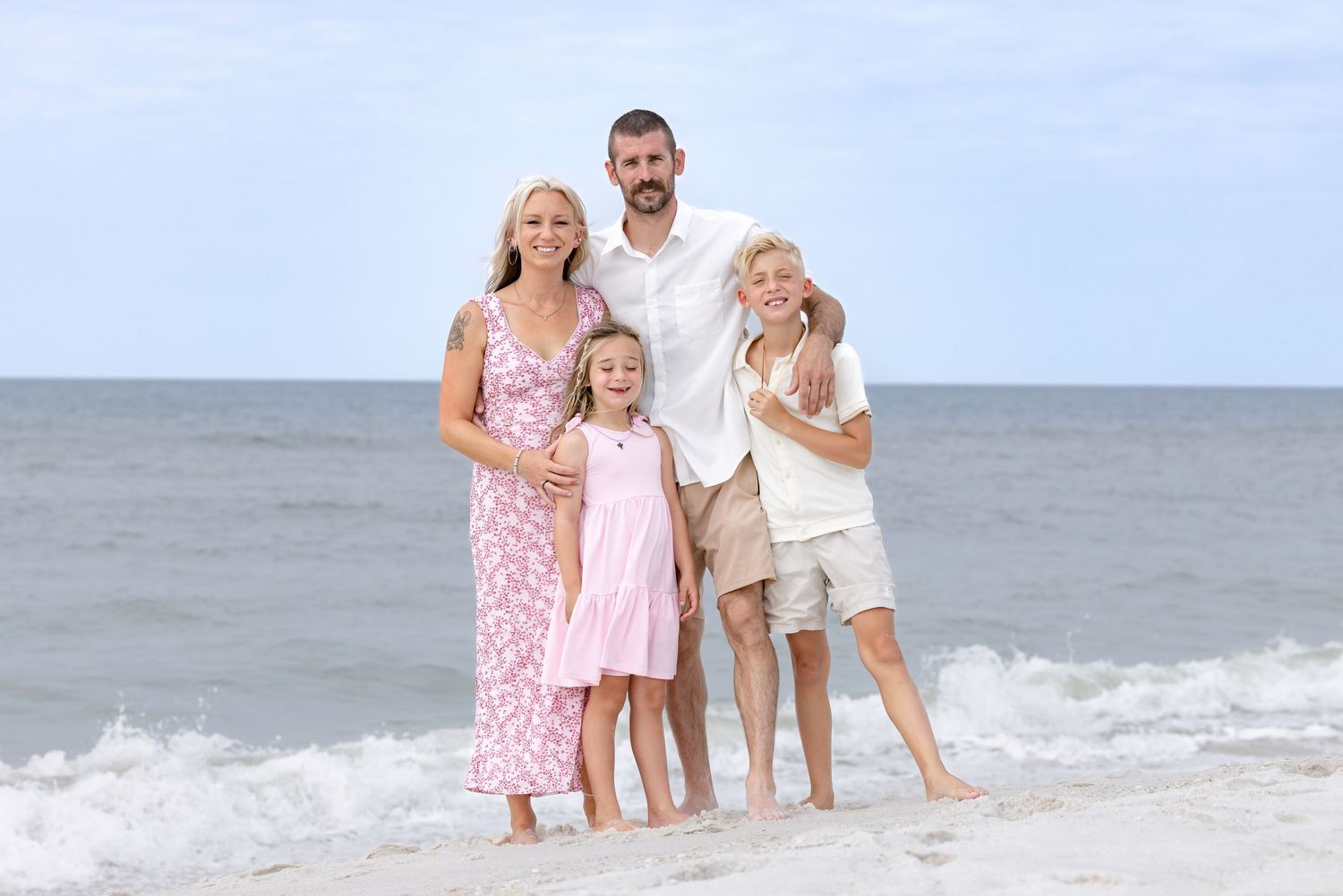 Family of four posing together on a sandy beach with ocean waves in the background on a sunny day.