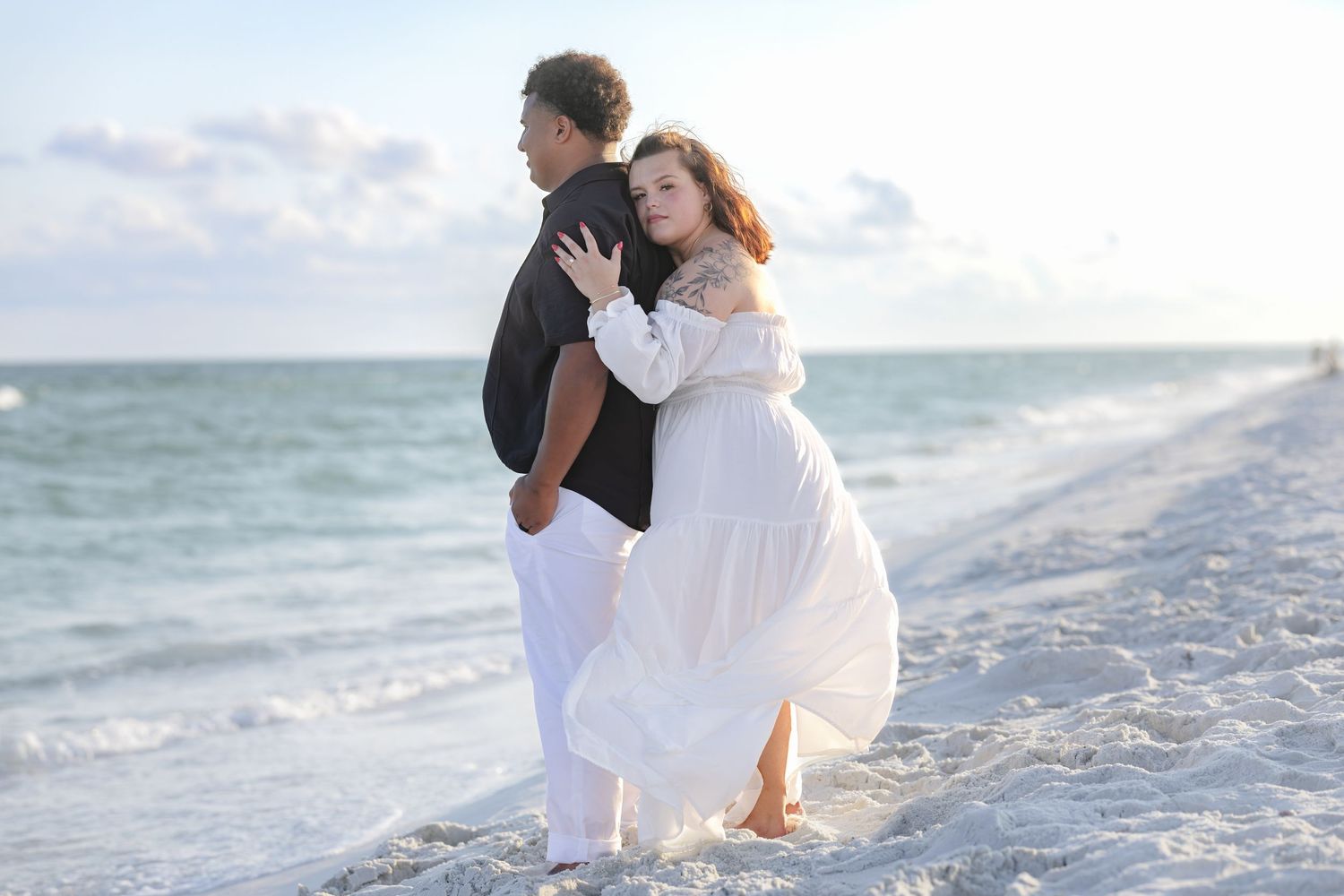 Romantic couple embracing on a sandy beach, woman in white dress and man in black shirt, ocean waves in background.