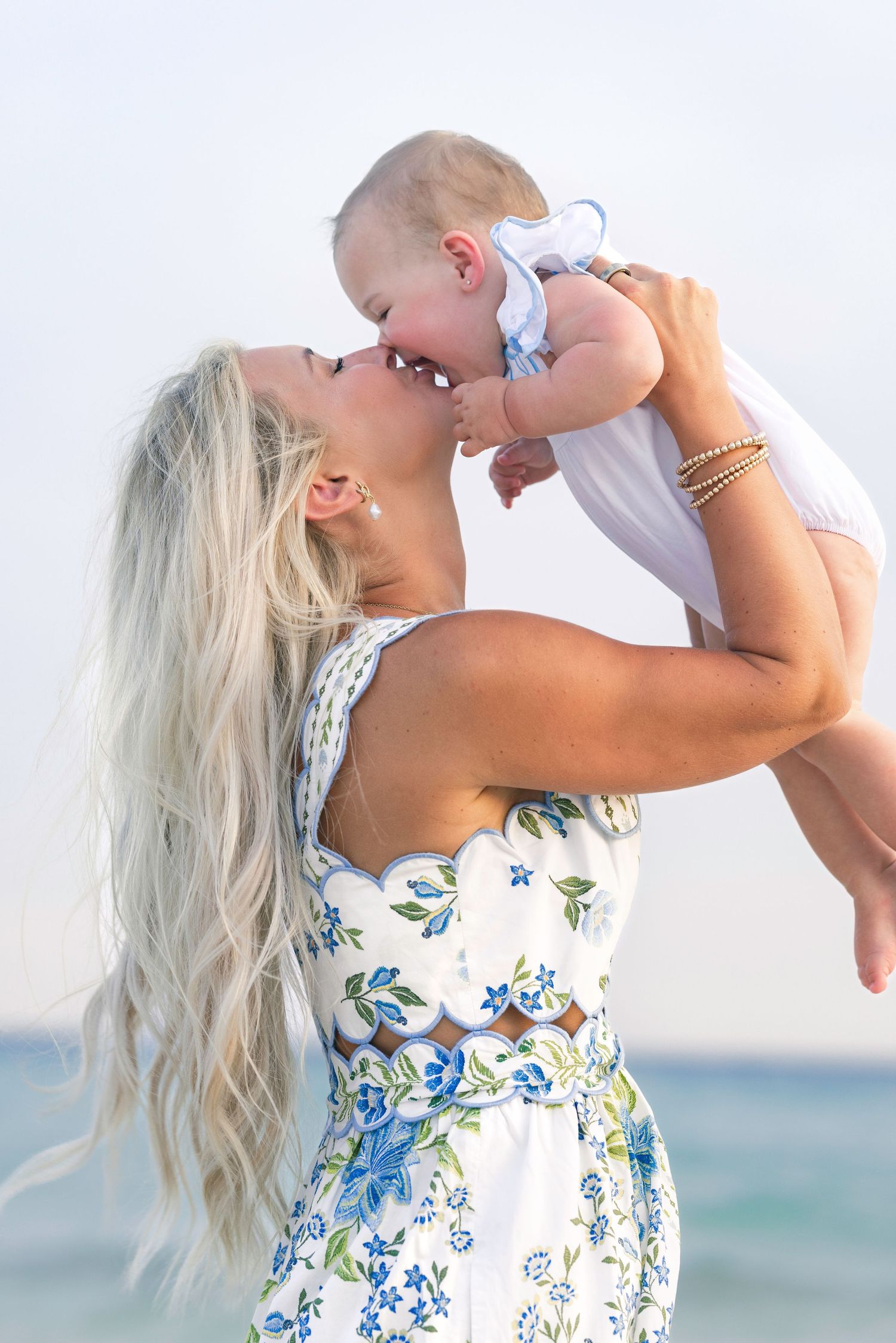 Blonde woman kissing her baby at the beach while lifting the infant up in the air.