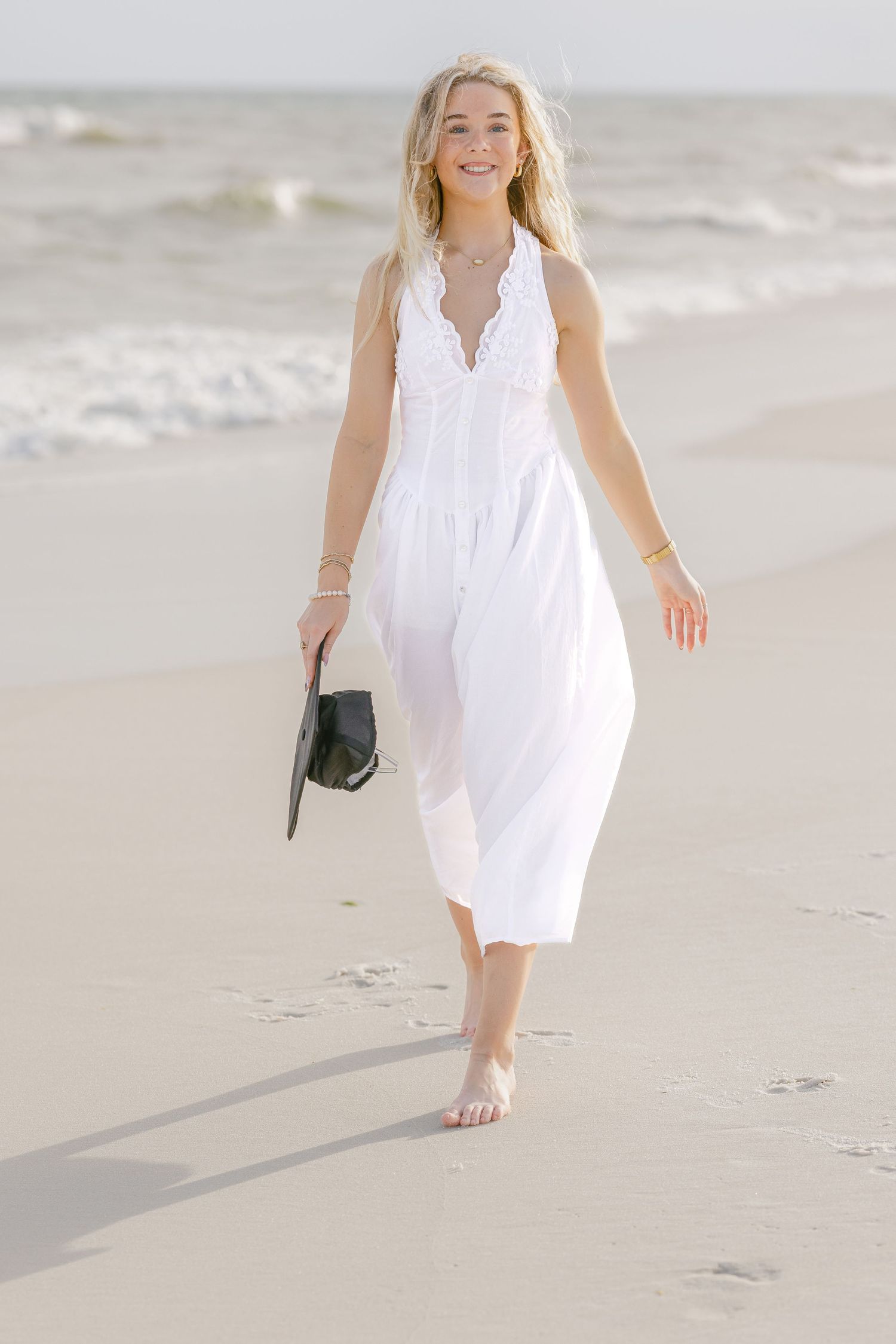 Smiling blonde woman in white lace maxi dress walking barefoot on sandy beach holding black sandals.