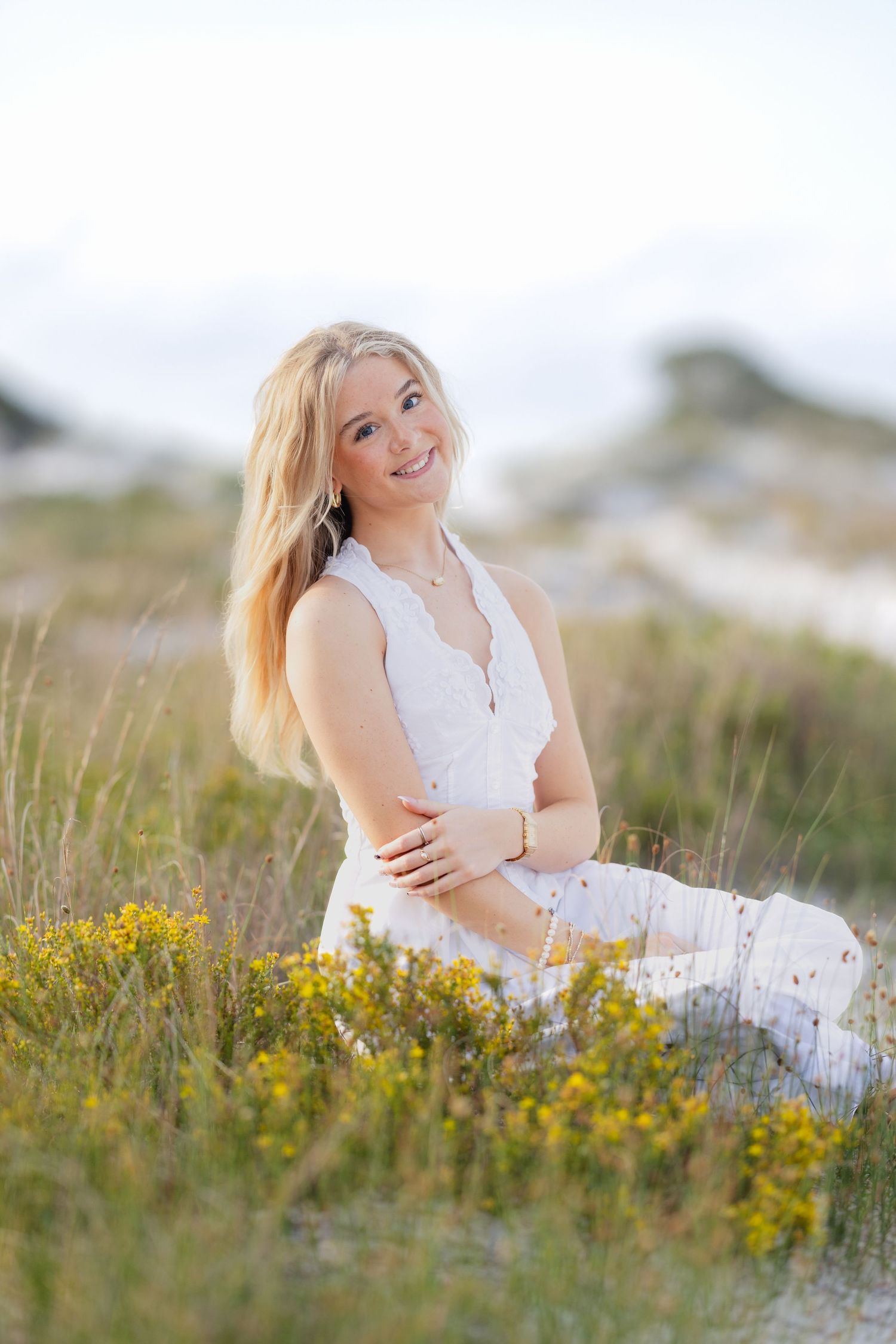 Smiling blonde woman in white dress sitting among wildflowers in a sunny field, portrait photography.