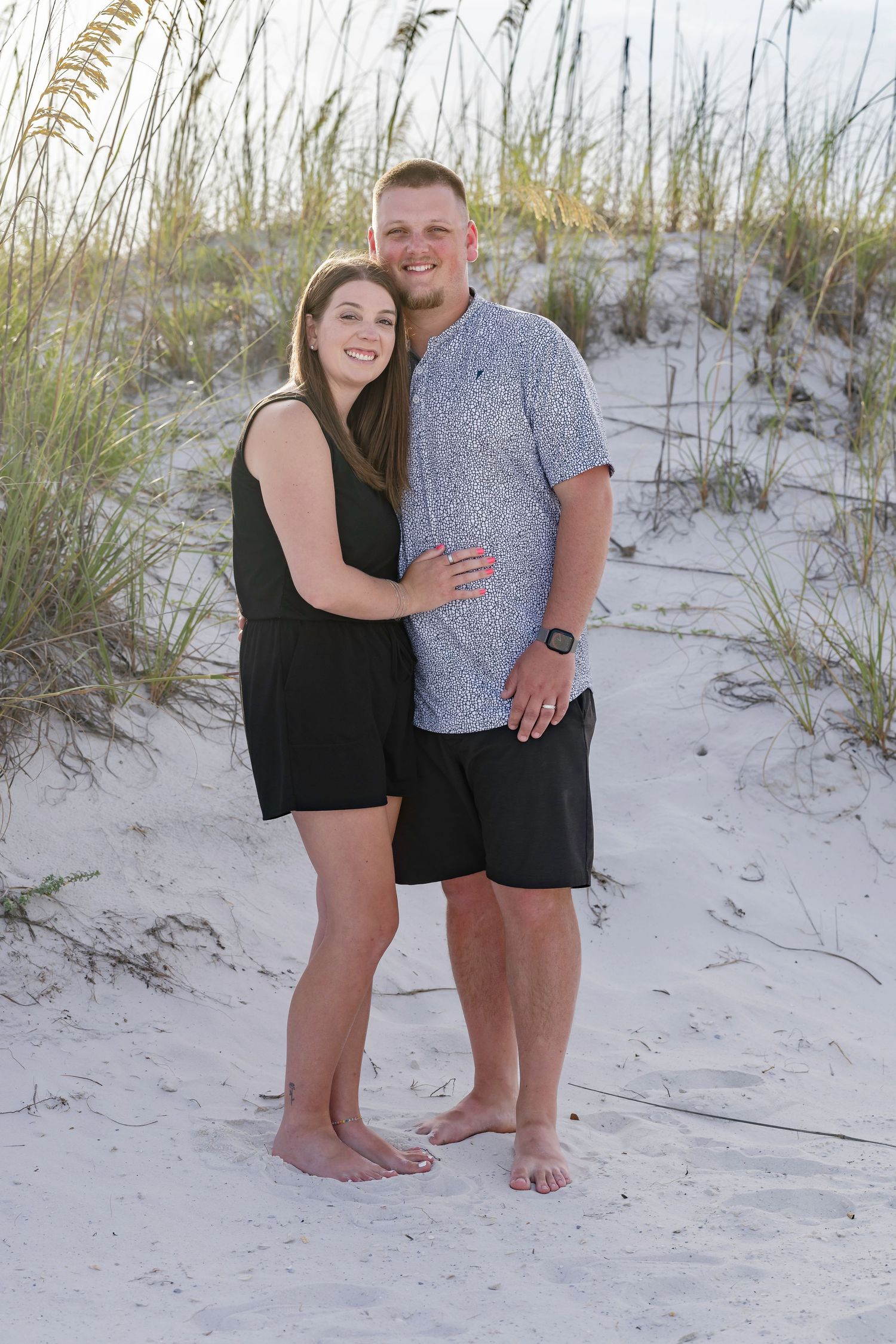 Couple smiling together on white sand beach dunes with sea oats, man in gray shirt and woman in black romper.