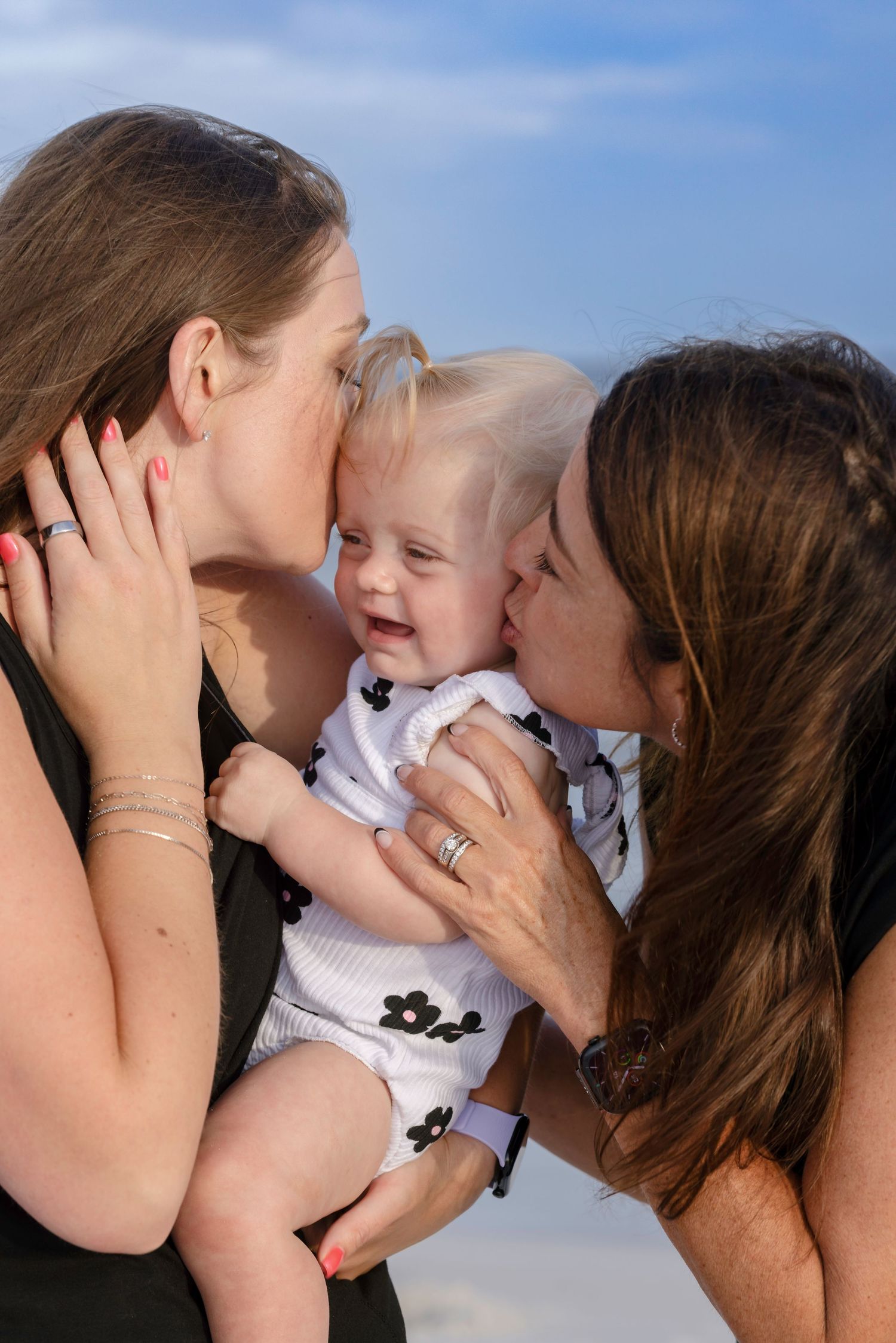 Two women kissing a happy laughing baby on the cheeks at a sunny beach.