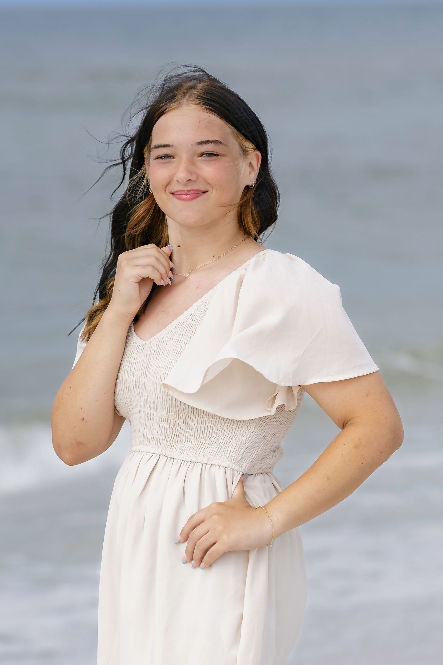Young woman in white ruffled dress smiling at beach with ocean waves in background.