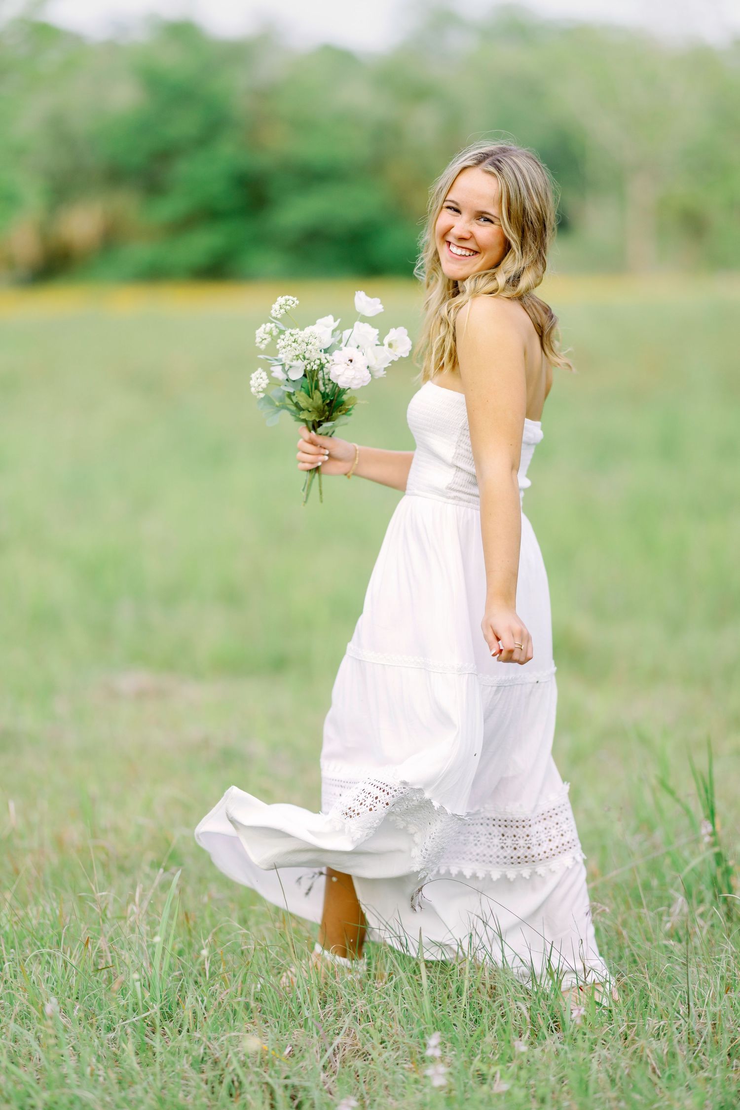 Person wearing a flowing white maxi dress holds white flowers while standing in a sunlit grassy field on a summer day.