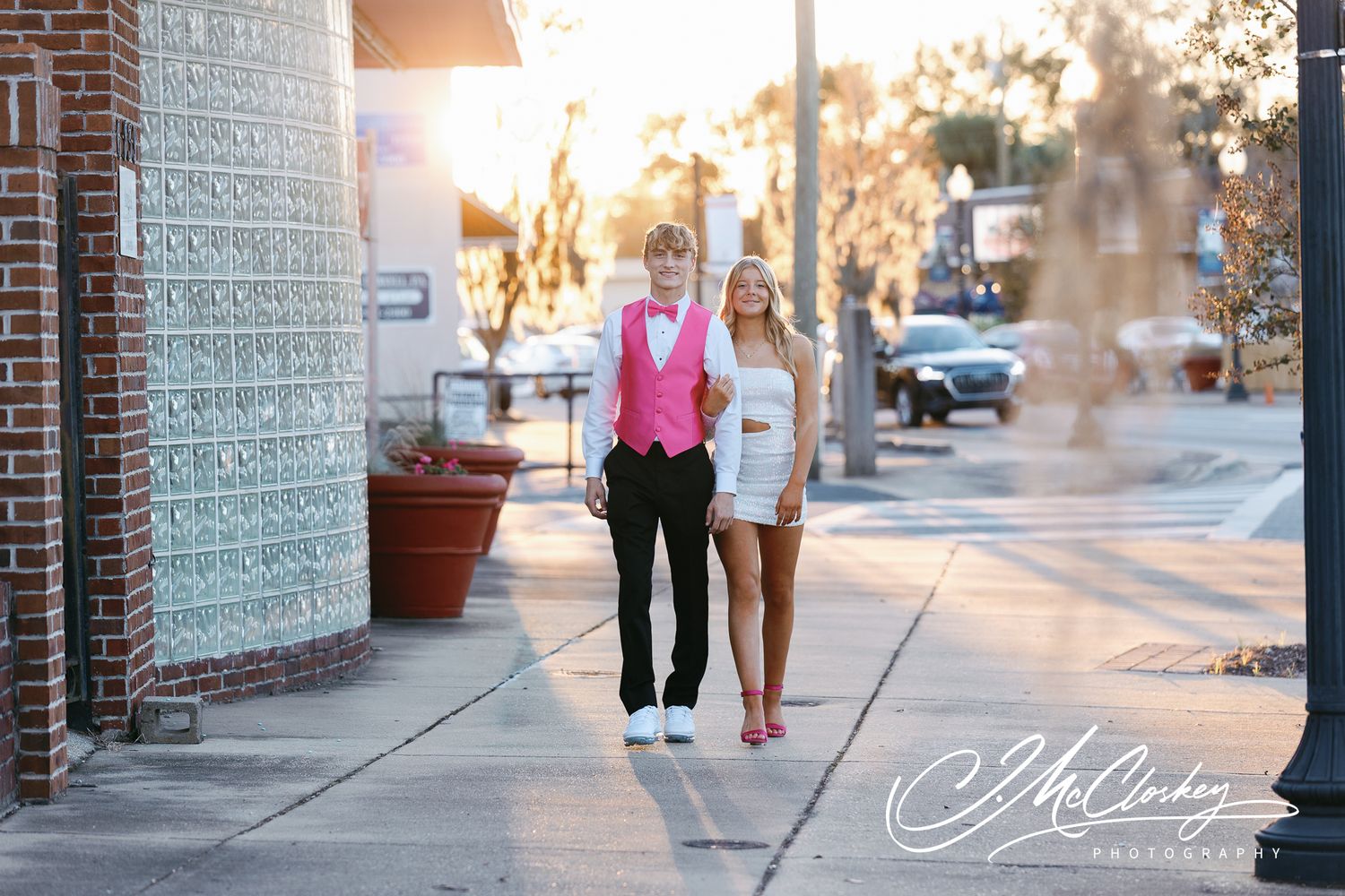 A couple walks together on a brick sidewalk during golden hour sunset with shadows casting on the pavement.