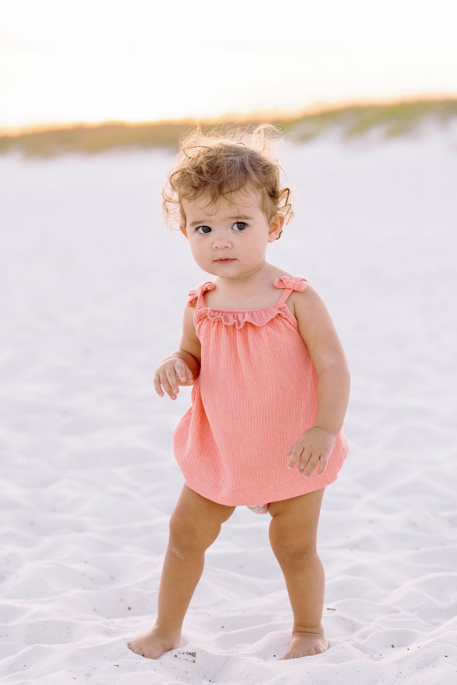 Baby wearing a coral-colored romper plays in white sand at the beach during golden hour.