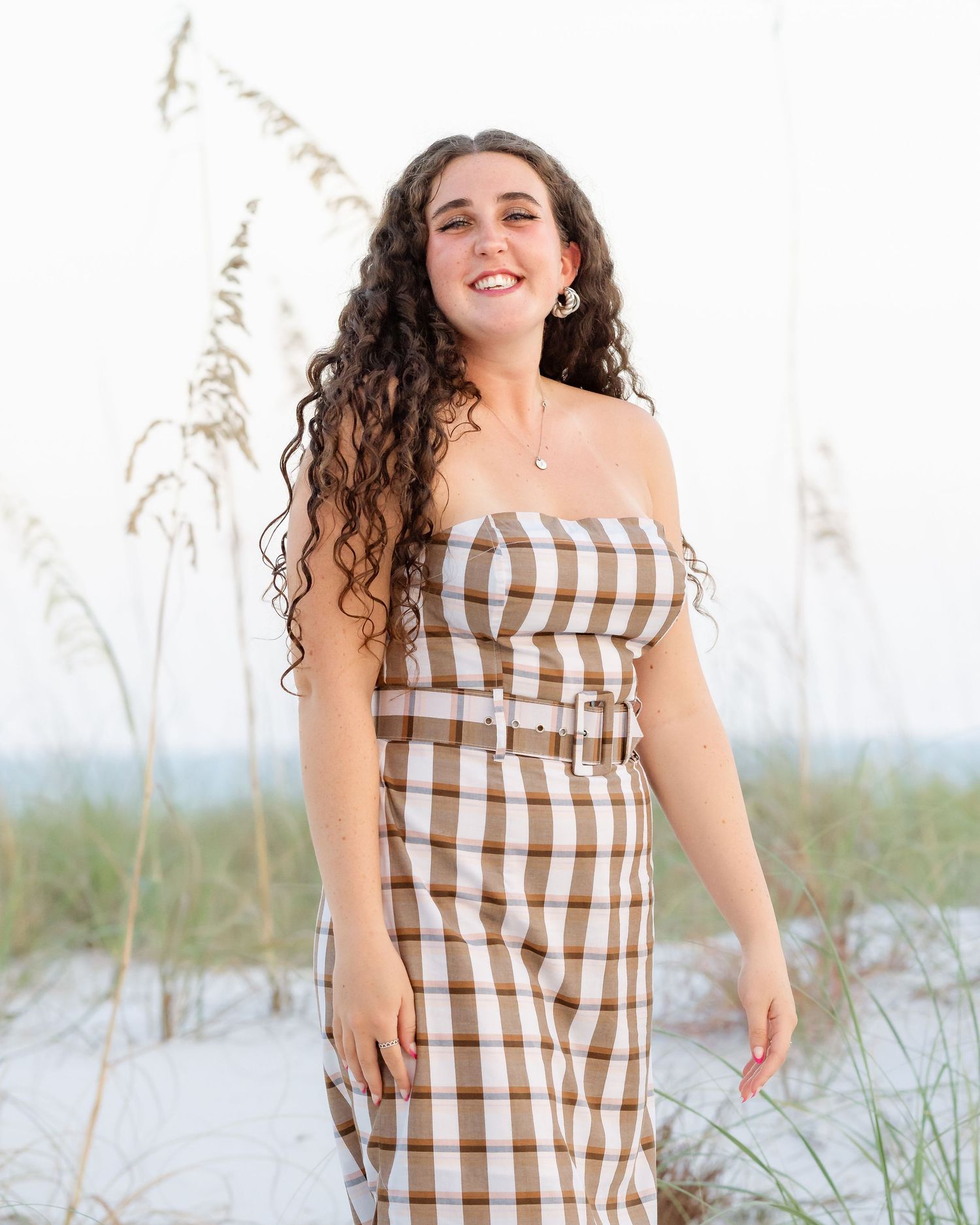 Young woman in brown plaid strapless belted dress smiling on sandy beach with sea oats in background.