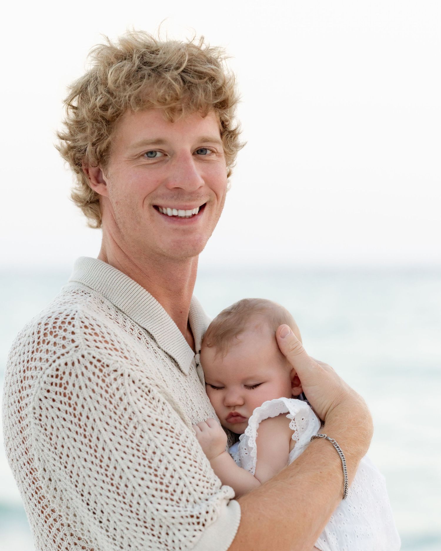 Smiling young man with curly blond hair holding a sleeping newborn baby on a sunny beach.