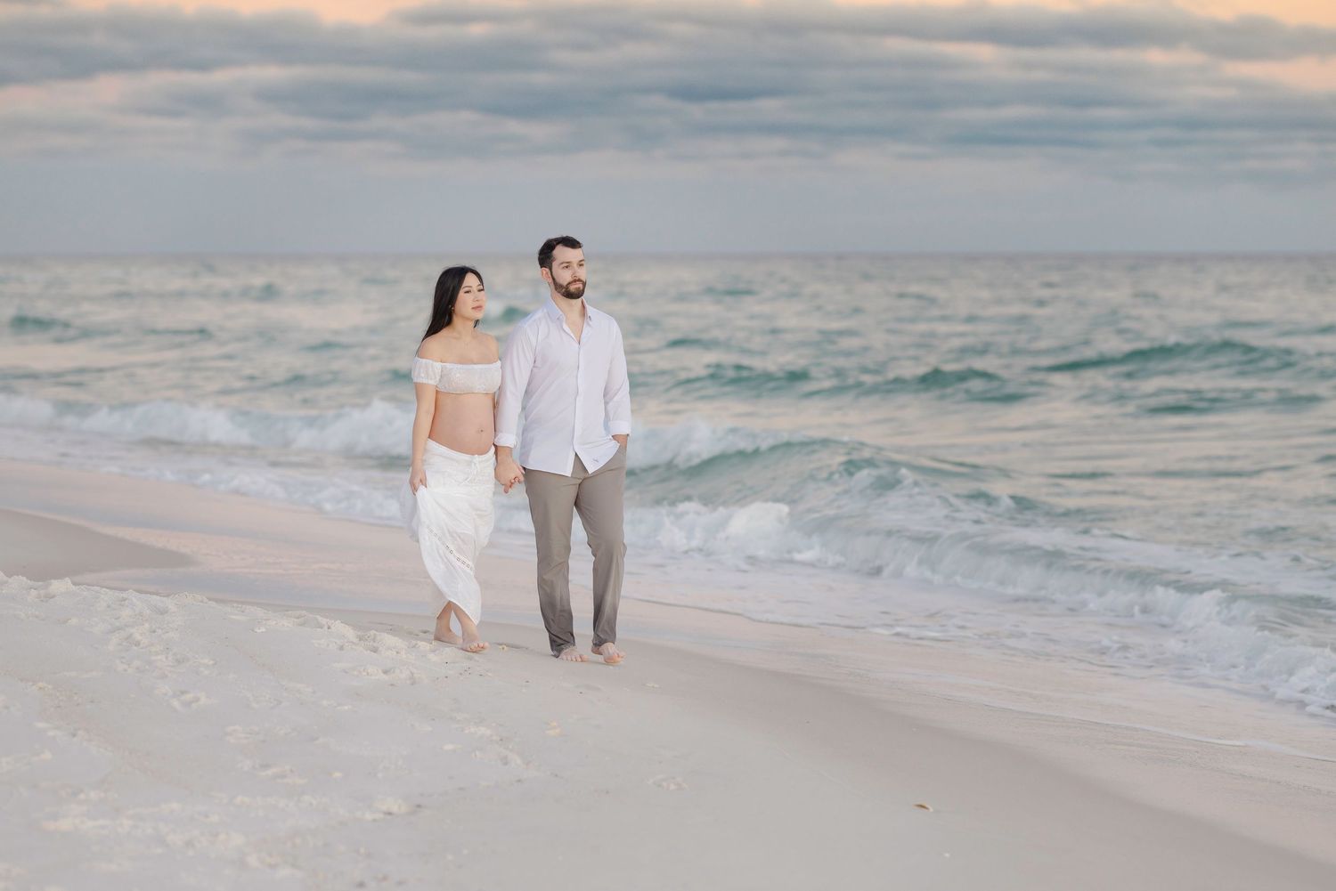 A couple walks hand in hand along a pristine beach at sunset with gentle waves rolling onto the shore.