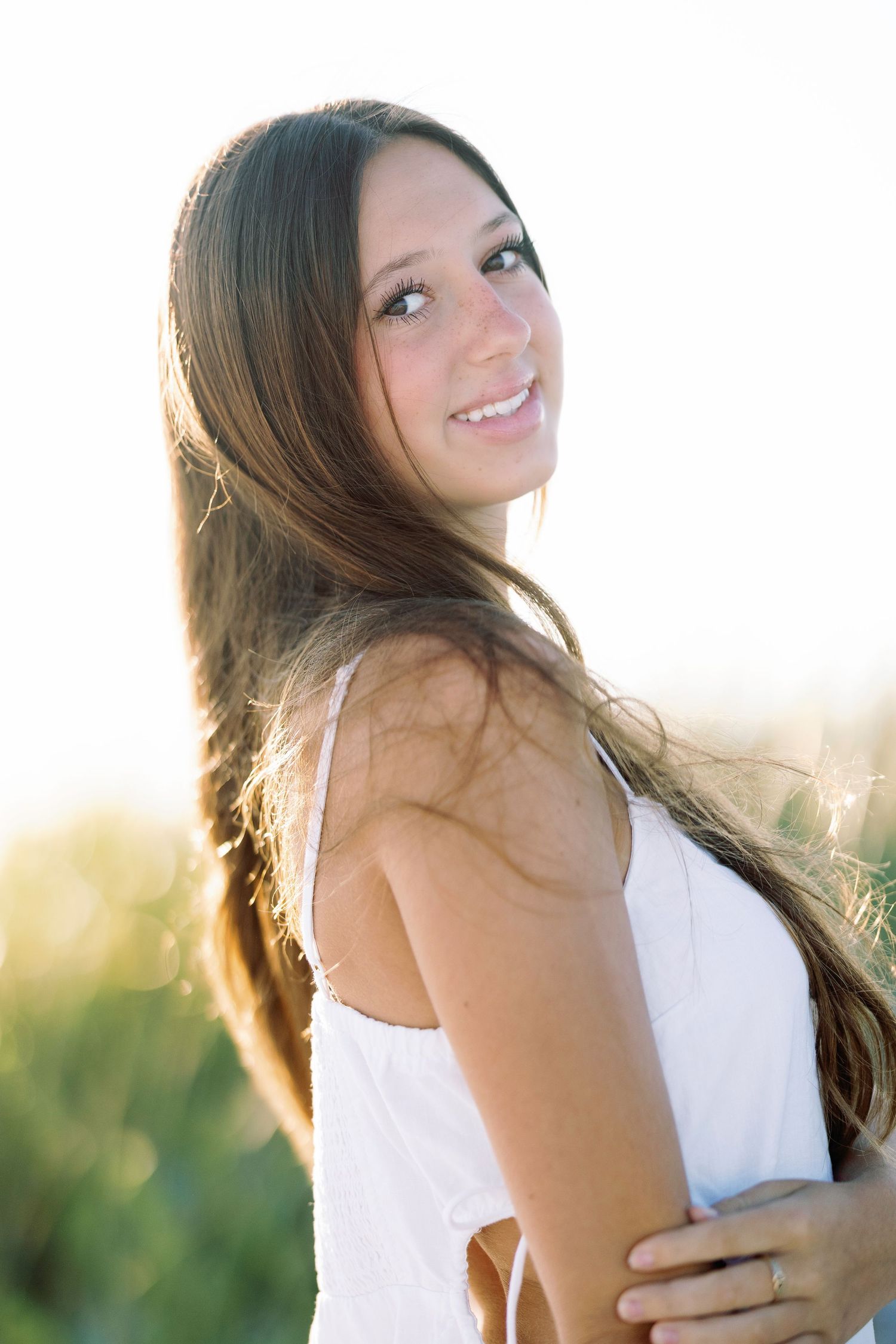 Woman in white dress poses against sunlit natural background with flowing hair.