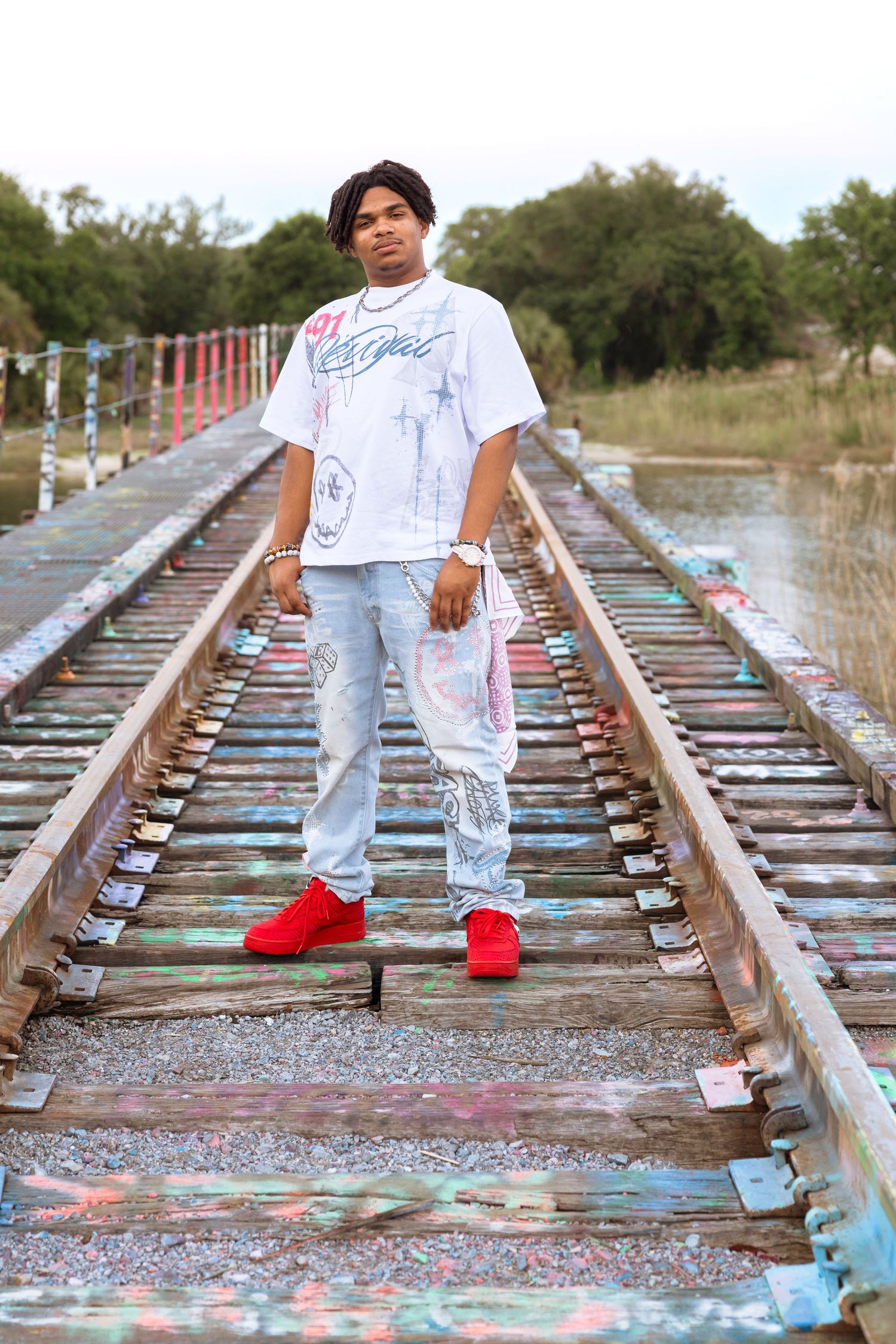 Young man in white graphic tee, light jeans, and red sneakers posing on graffiti-covered railroad tracks outdoors.