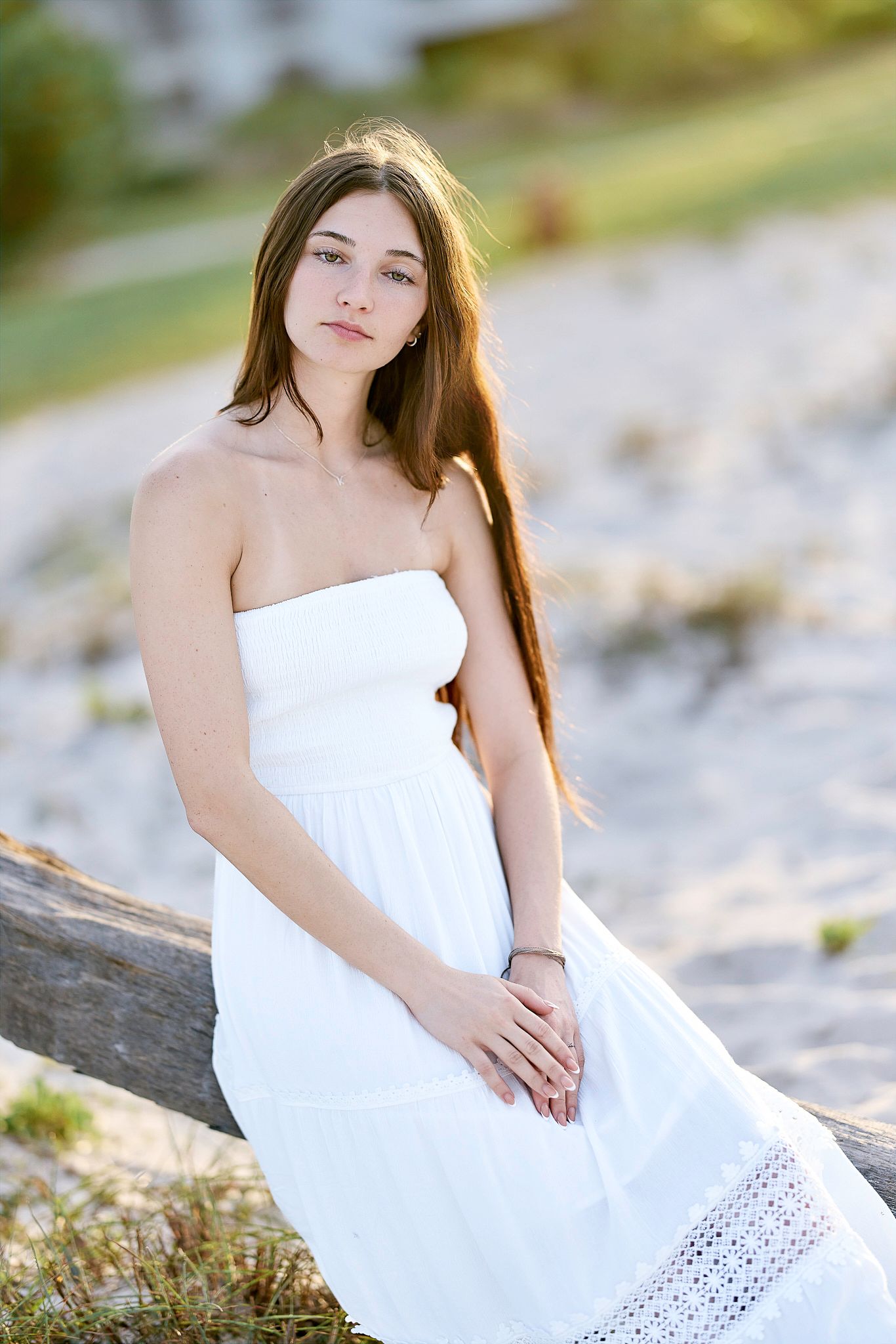 A woman in a white sundress poses outdoors in soft natural lighting near a rocky beach area.