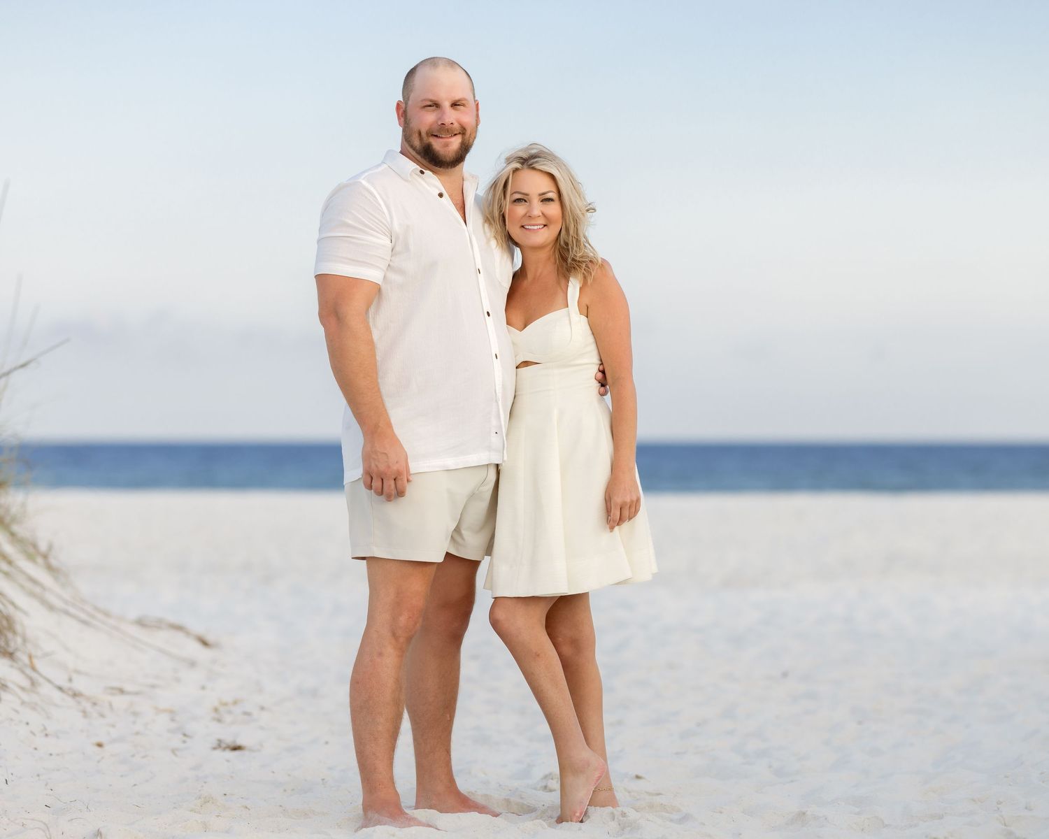 A couple in matching white outfits poses barefoot on a sandy beach with calm ocean waters in the background.