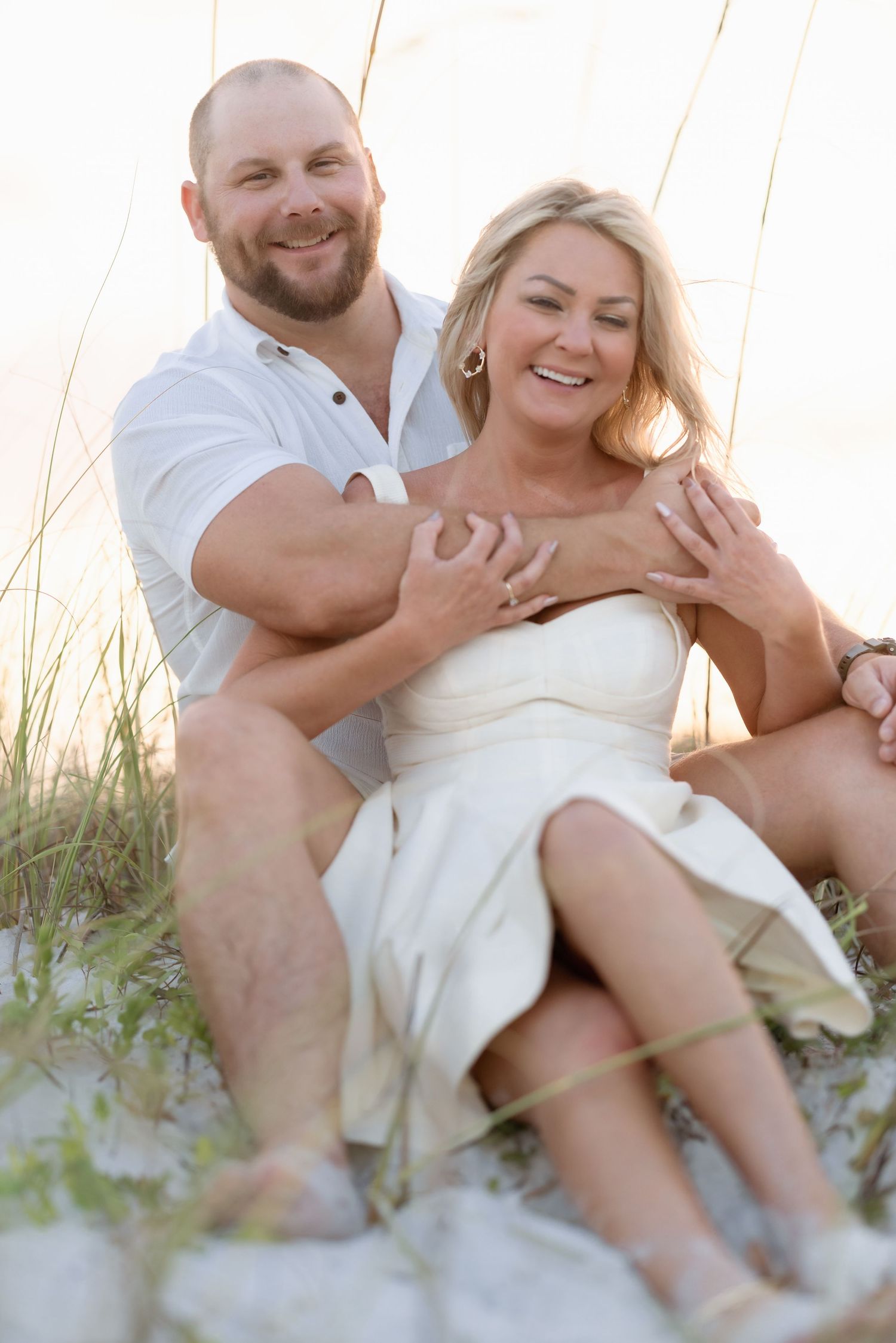 Happy couple sitting together on sandy beach dunes at sunset, woman in white dress, man in white shirt.