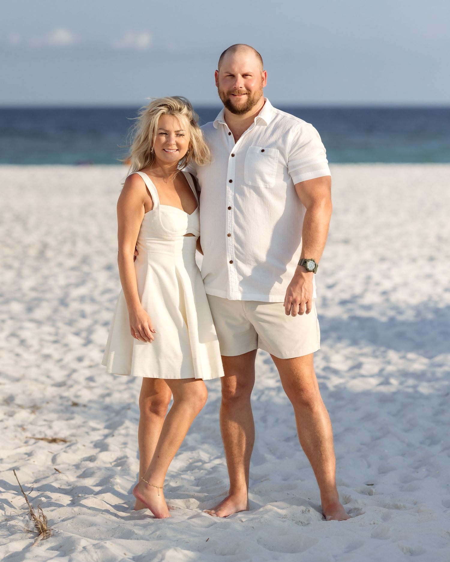 Couple posing together on a white sand beach, both wearing white outfits, smiling with ocean in the background.